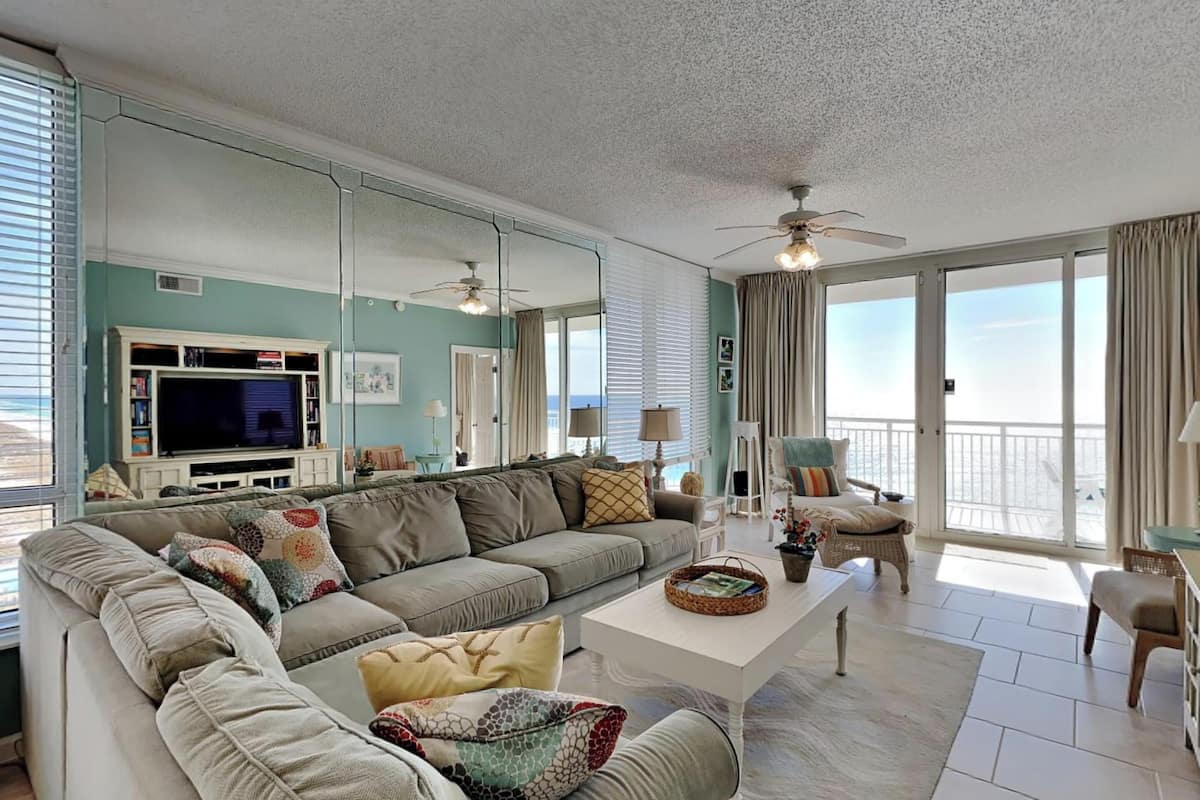A spacious living room with a large sectional sofa, a flat-screen TV, and a balcony with a view of the ocean.