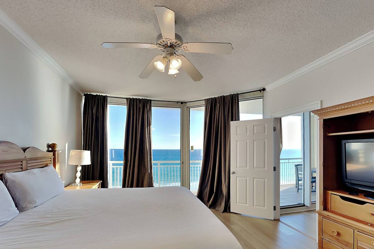A hotel room with a large bed, a ceiling fan, a TV, a wooden dresser, and a balcony with a view of the ocean.