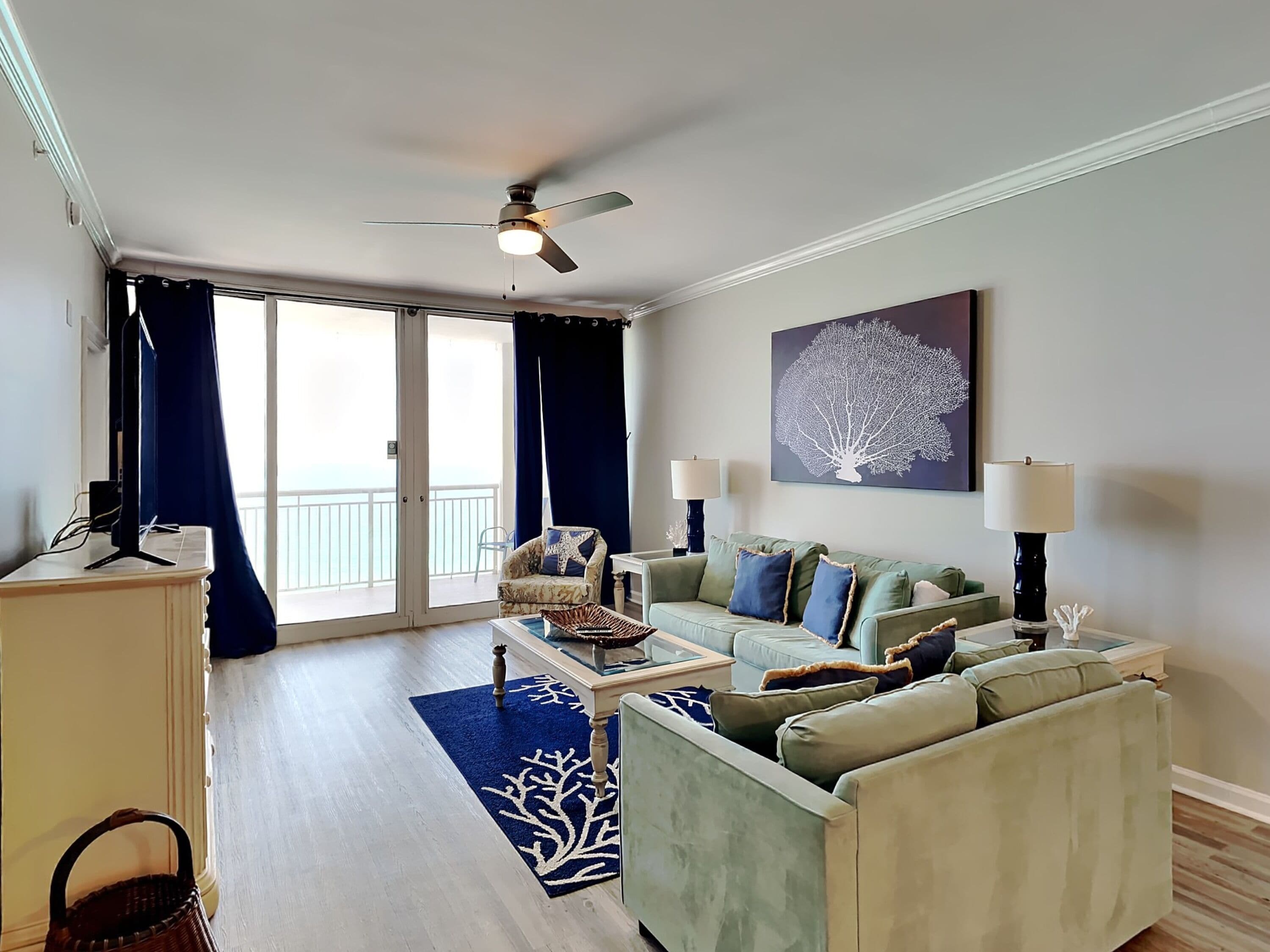 Ein Wohnzimmer mit einer Couch, einem Couchtisch und einem Deckenventilator. Ein großes Fenster mit Vorhängen bietet Blick ins Freie.