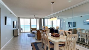 En rummelig opholdsstue med en stor glasvæg, der giver udsigt til havet. Der er et spisebord med stole, en sofa og et fjernsyn monteret på væggen.