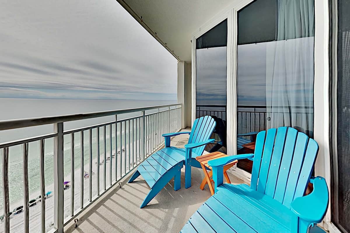 A balcony with two blue Adirondack chairs and a small table, overlooking a beach with a view of the ocean.