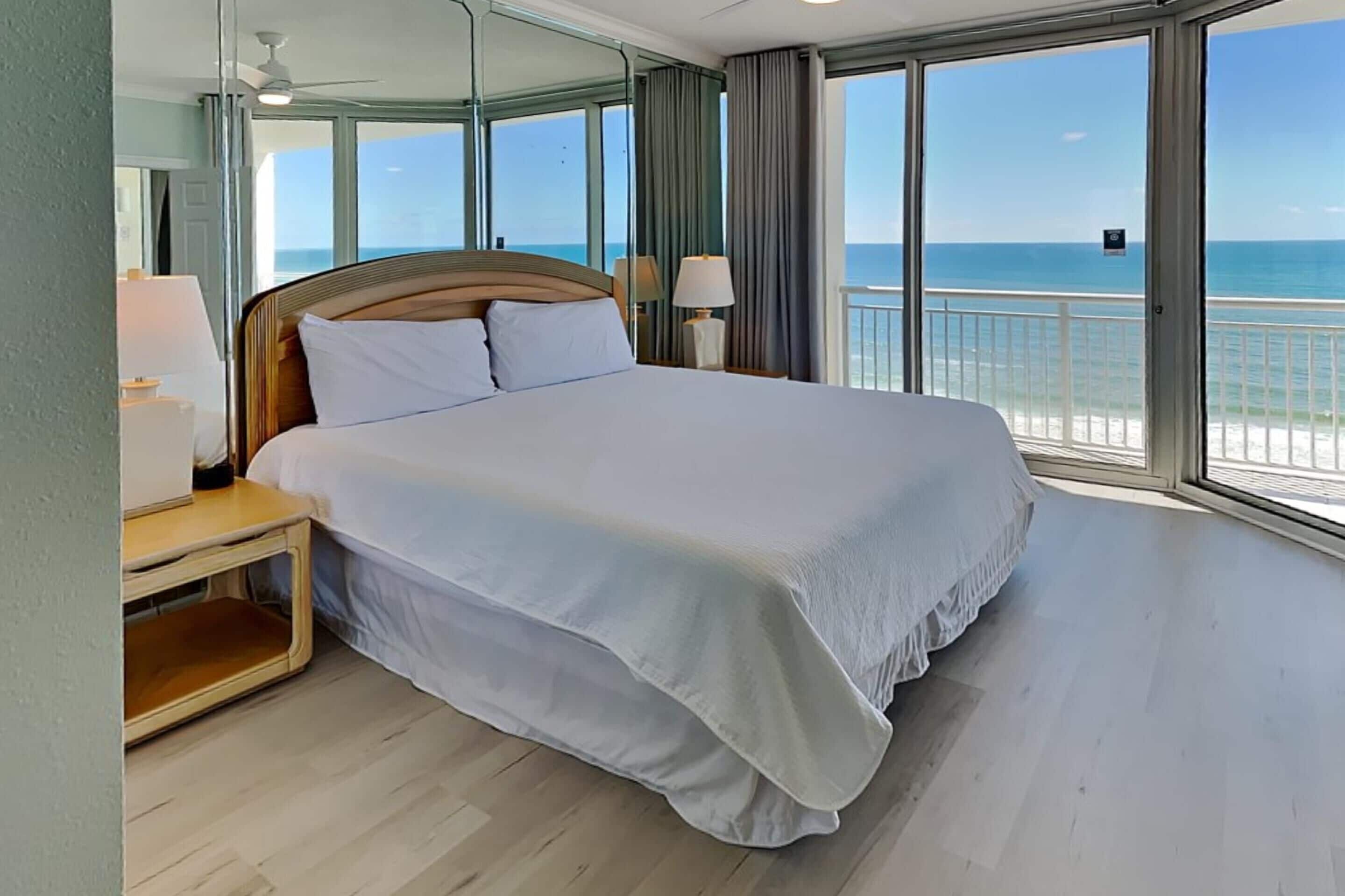 Una cama grande con sábanas blancas, un cabecero de madera y una mesita de noche con una lámpara. La habitación tiene ventanas amplias con vistas al océano.