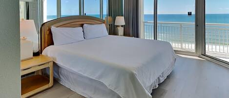 Una cama grande con sábanas blancas, un cabecero de madera y una mesita de noche con una lámpara. La habitación tiene ventanas amplias con vistas al océano.