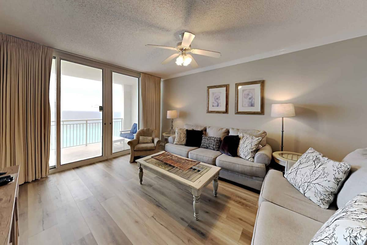 A spacious living room with a large sliding glass door leading to a balcony, a ceiling fan, a sofa set, and two framed artworks on the wall.