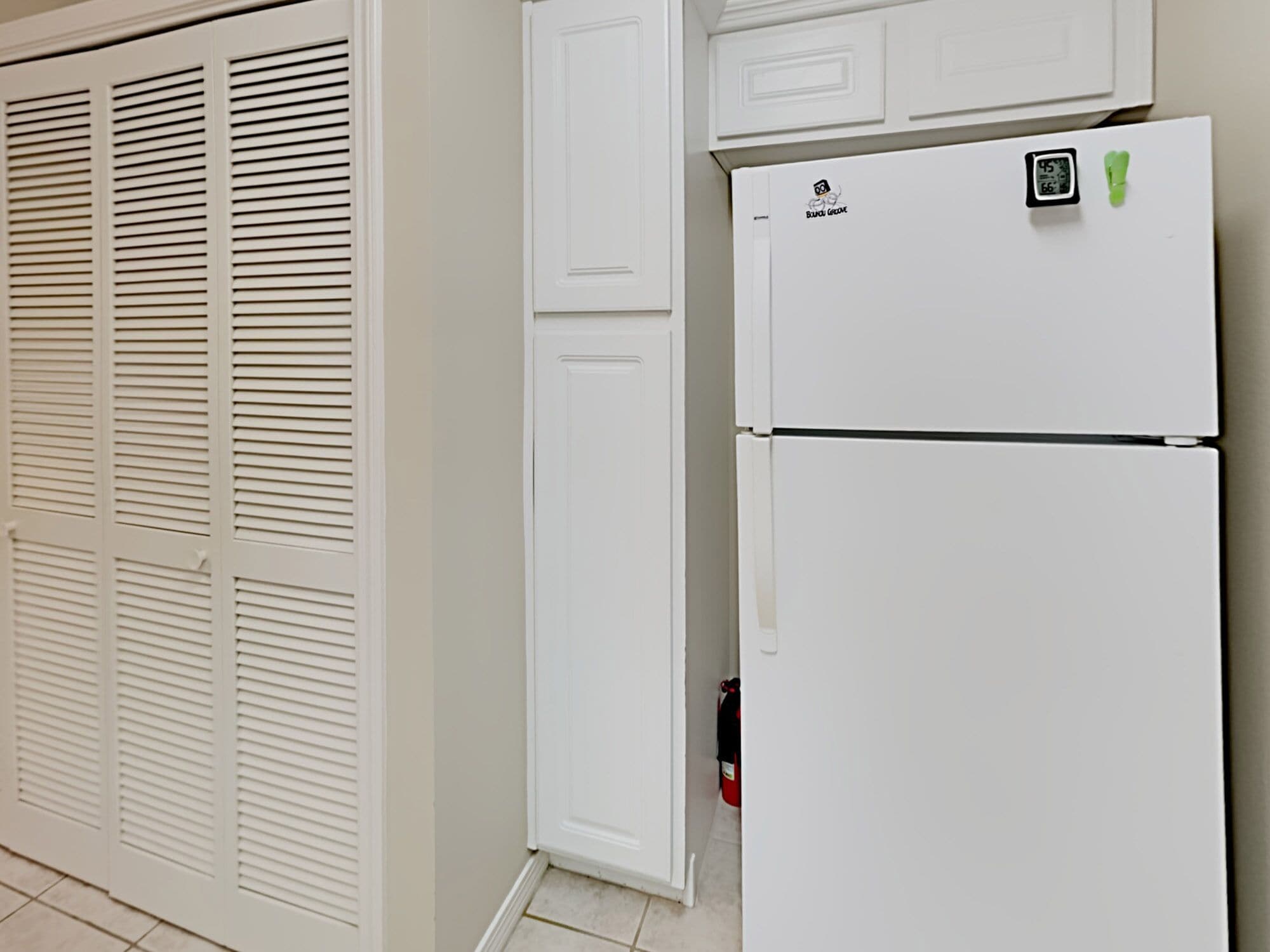 Un réfrigérateur blanc doté d’un écran numérique et d’une pince verte, placé à côté d’une armoire blanche et d’une porte blanche ornée de panneaux à persiennes horizontales.
