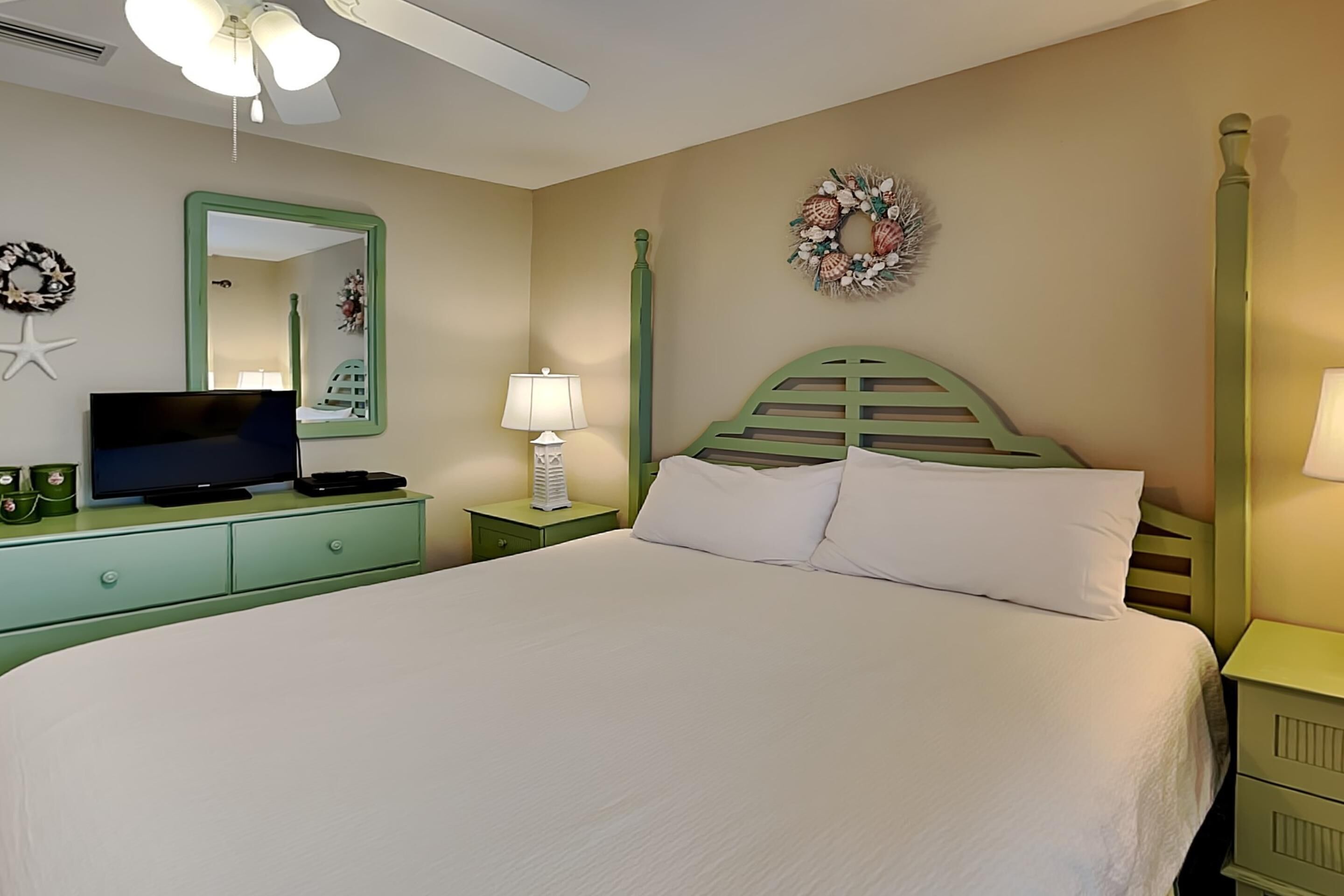 A neatly arranged bedroom with a bed, a television on a dresser, a mirror, and a wall decoration.