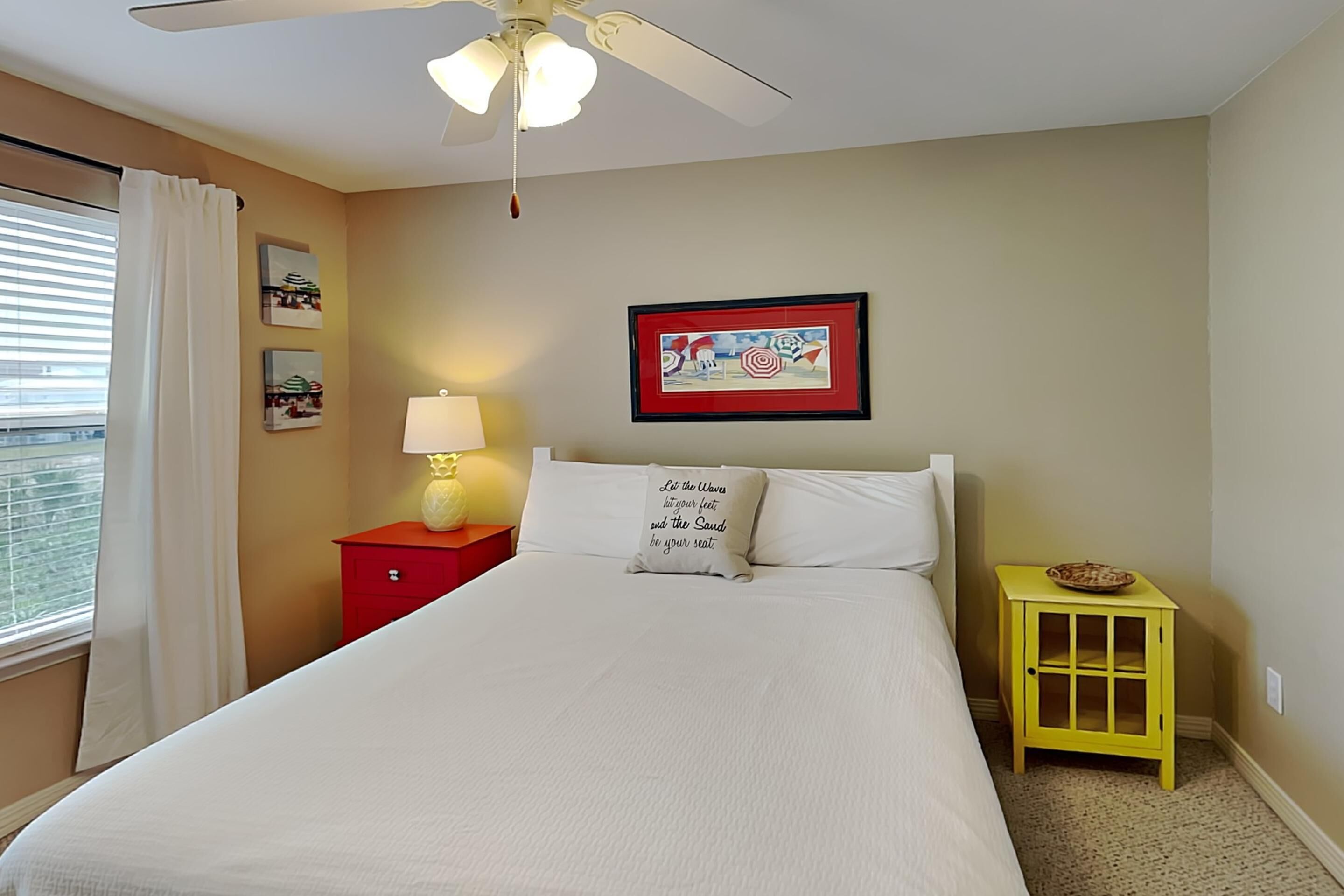 A bedroom with a bed, a nightstand, a lamp, a ceiling fan, a window with curtains, and a framed picture on the wall.