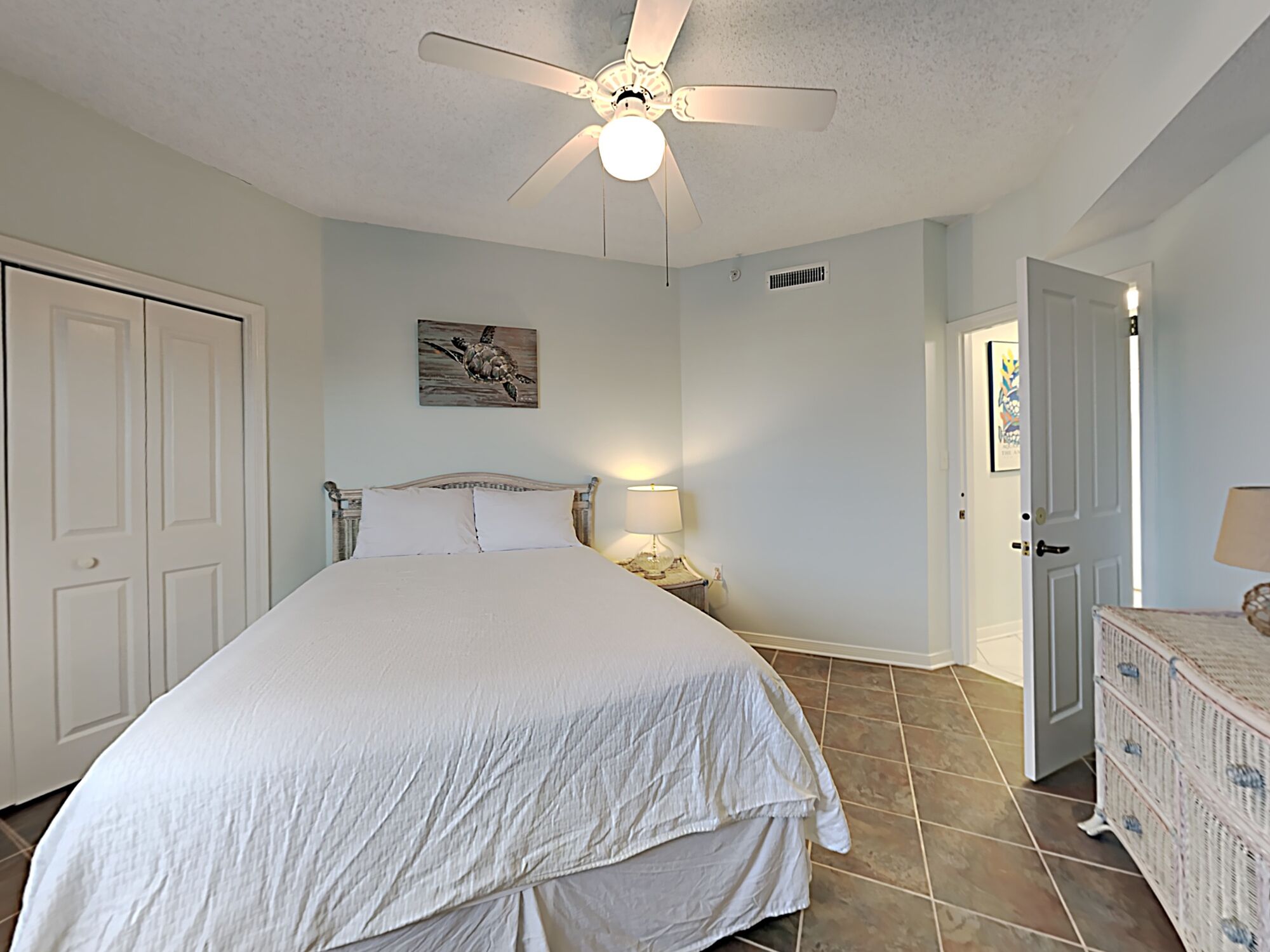 A bedroom with a large bed, a ceiling fan, a nightstand with a lamp, and a dresser.