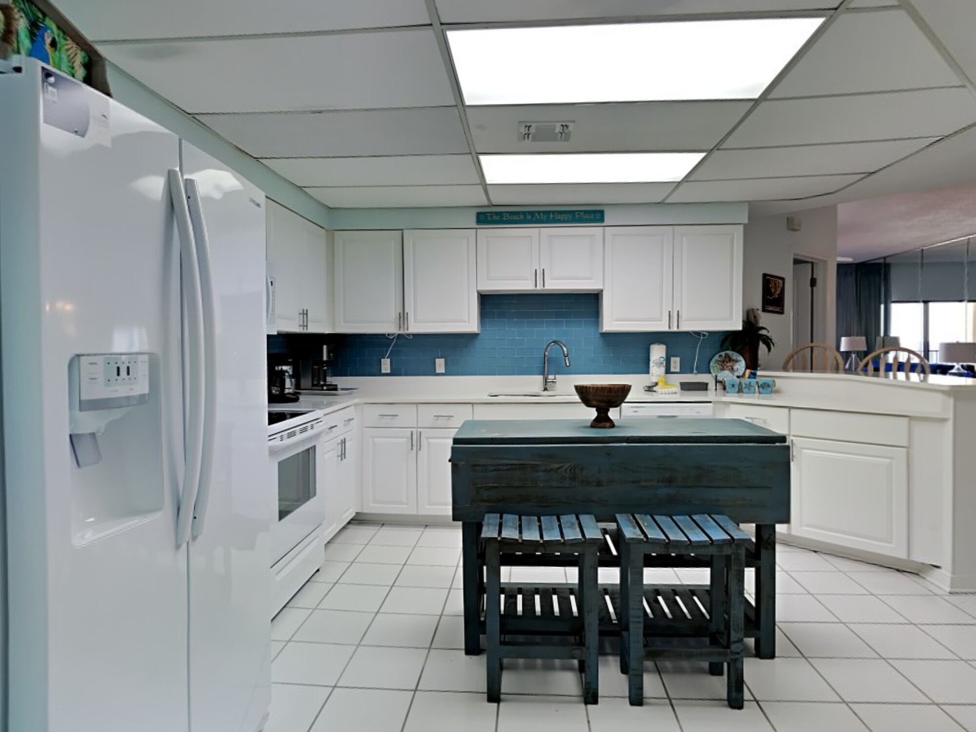 Una cocina moderna con armarios blancos, salpicadero azul, una isla central con taburetes y un refrigerador a la izquierda.