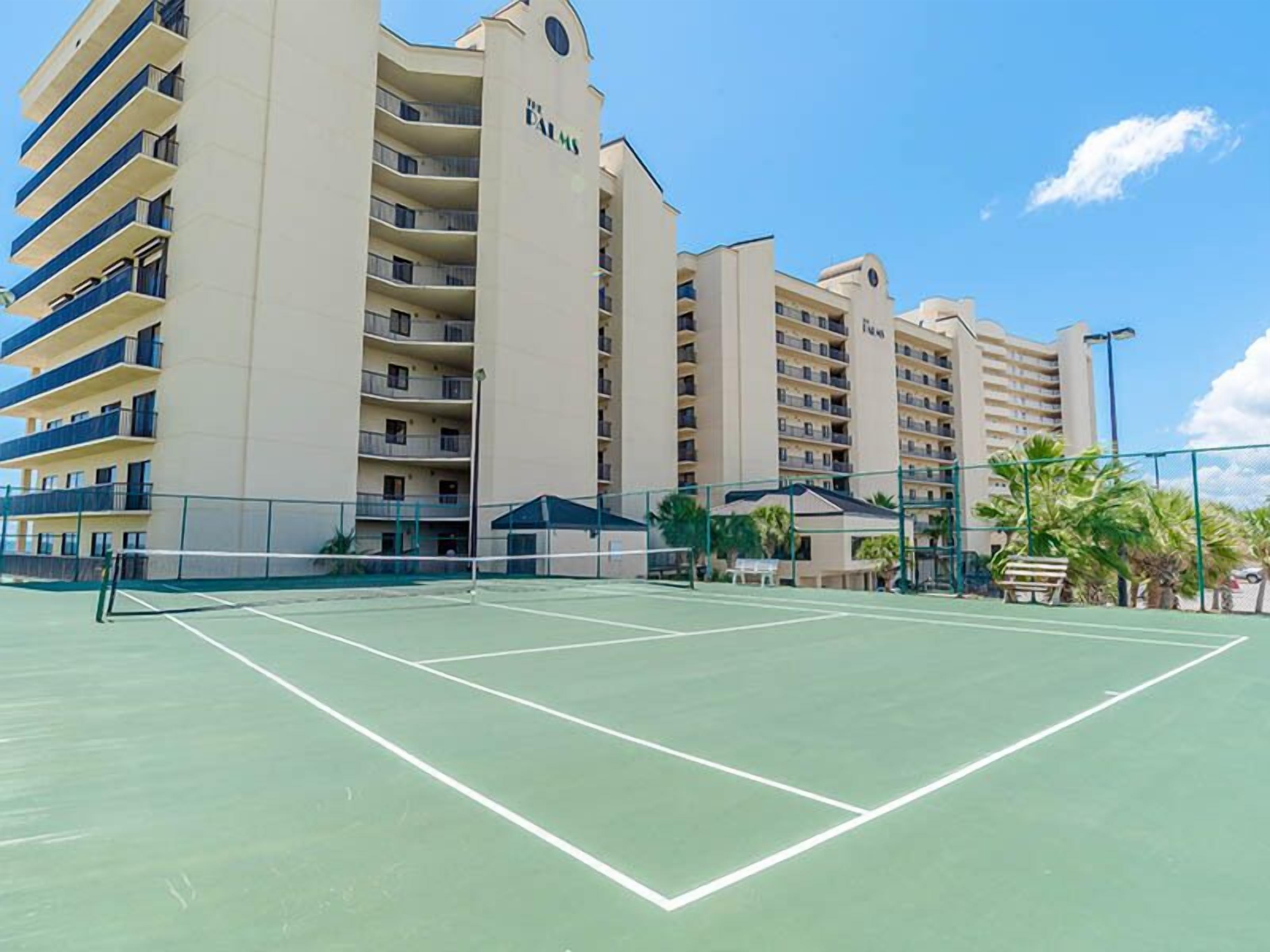 A tennis court with a net in the middle, surrounded by a fence. There are palm trees and a building with balconies in the background.