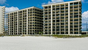 Un hotel di grandi dimensioni con numerosi piani e balconi, affacciato su una spiaggia sabbiosa sotto un cielo sereno.