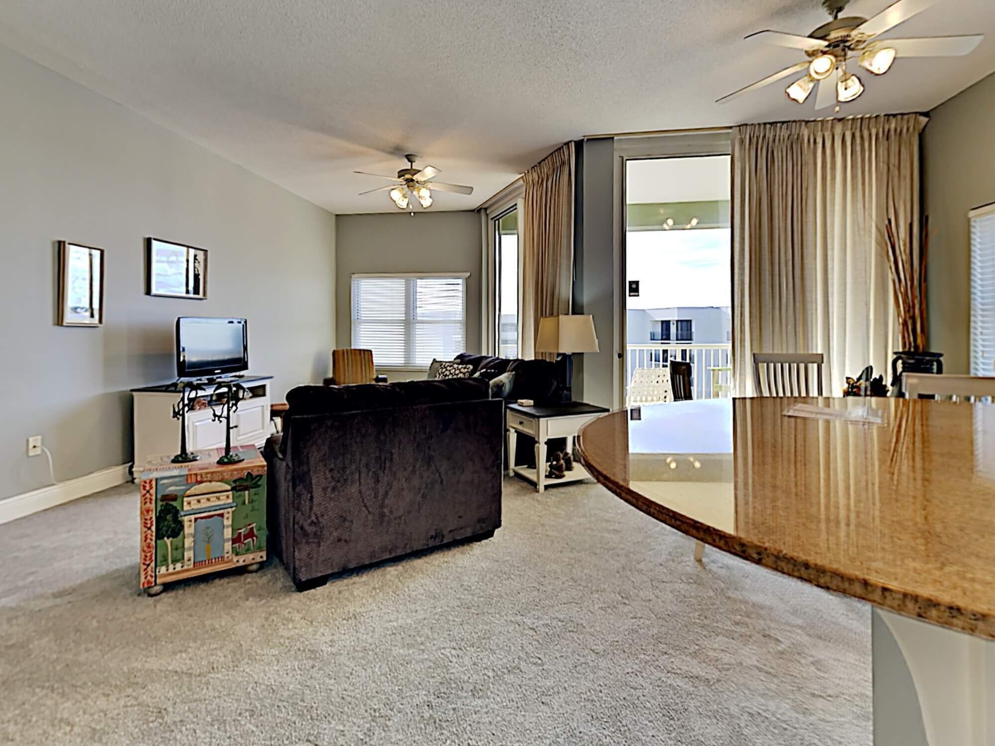 A spacious living area with a sofa, a television, and a small table. There is a balcony with a view of the outside.