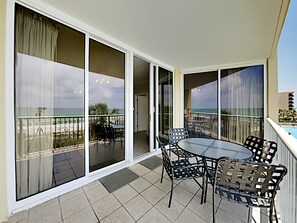 A balcony with a glass door leading to a terrace with a table and chairs, overlooking a beach. - Water's Edge by Southern Vacation Rentals (Fort Walton Beach)