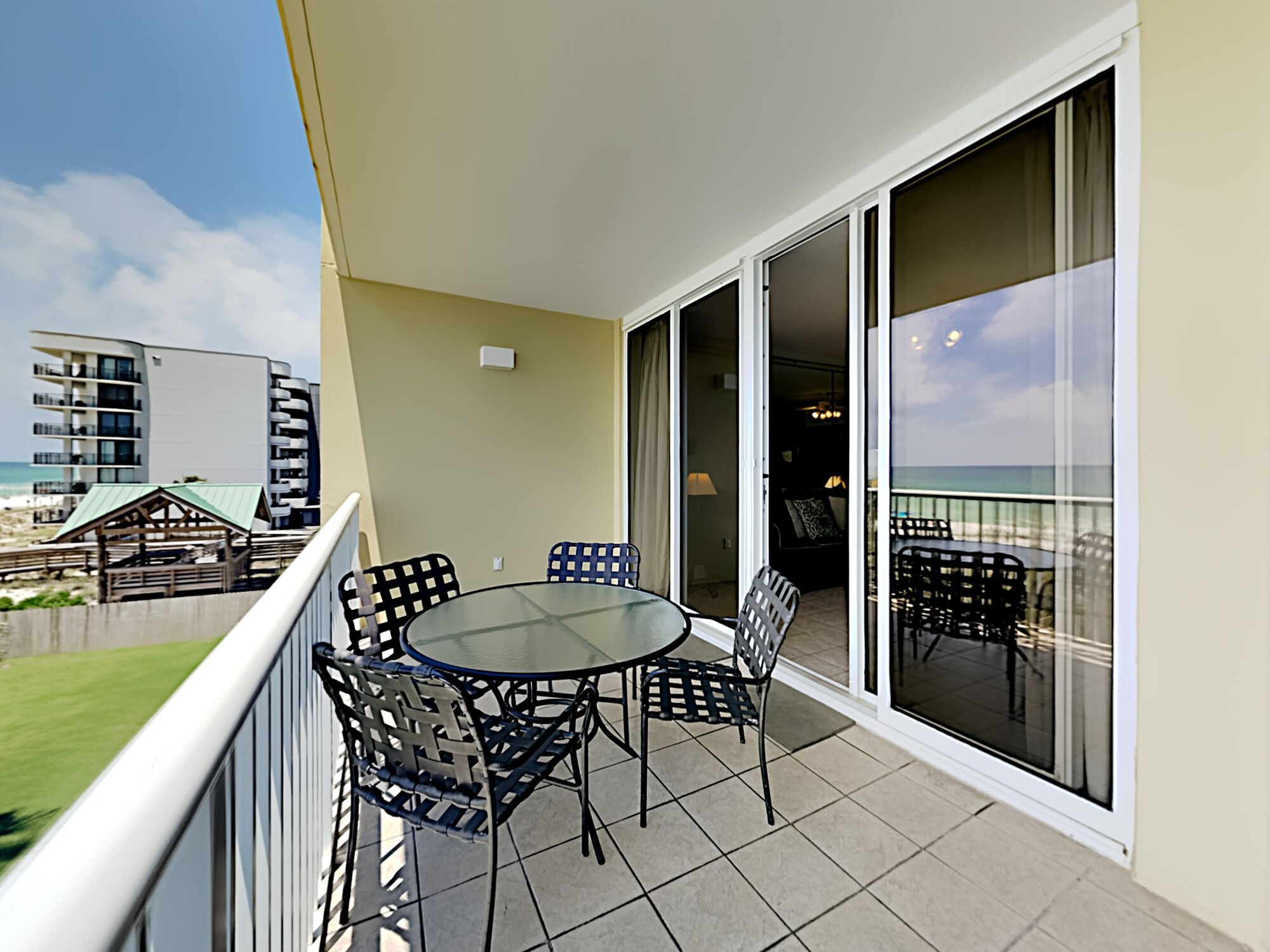 Een balkon met een glazen tafel en stoelen, uitzicht op het strand en een gebouw op de achtergrond.