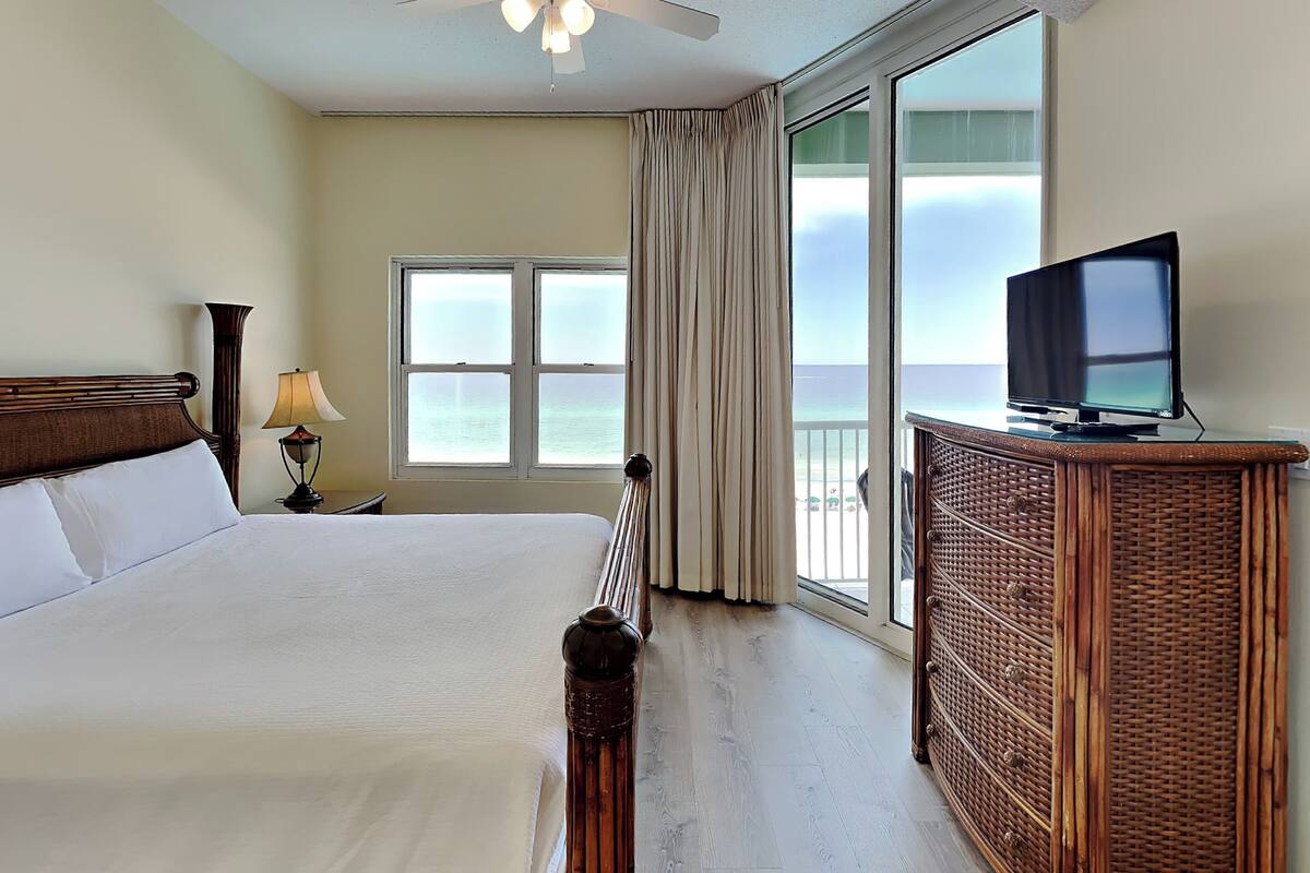 Une chambre d’hôtel avec un lit, un téléviseur posé sur une commode en bois, un ventilateur de plafond et une grande fenêtre donnant sur l’océan.