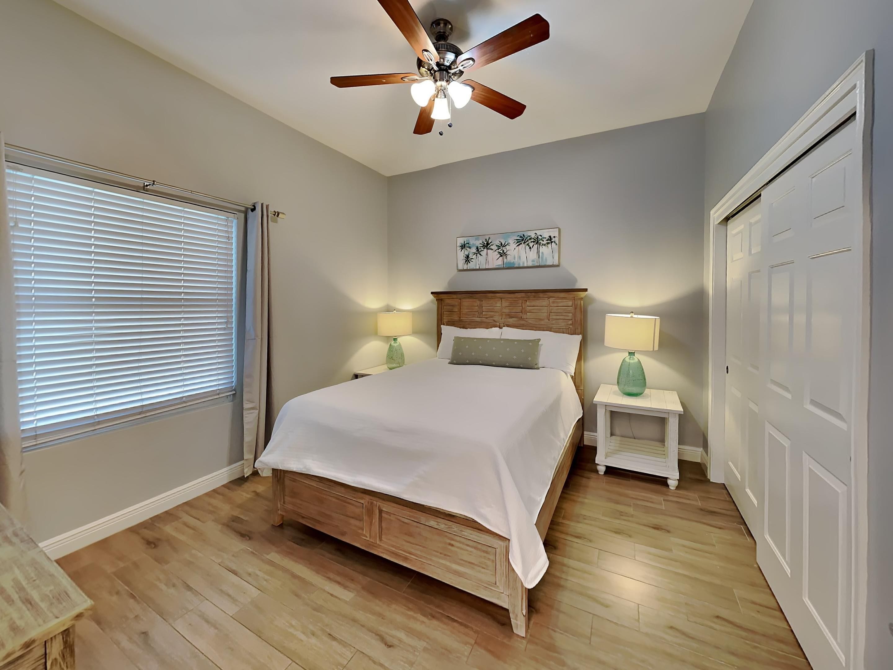 A bedroom with a wooden bed, two bedside tables with lamps, a ceiling fan, and a window with blinds.
