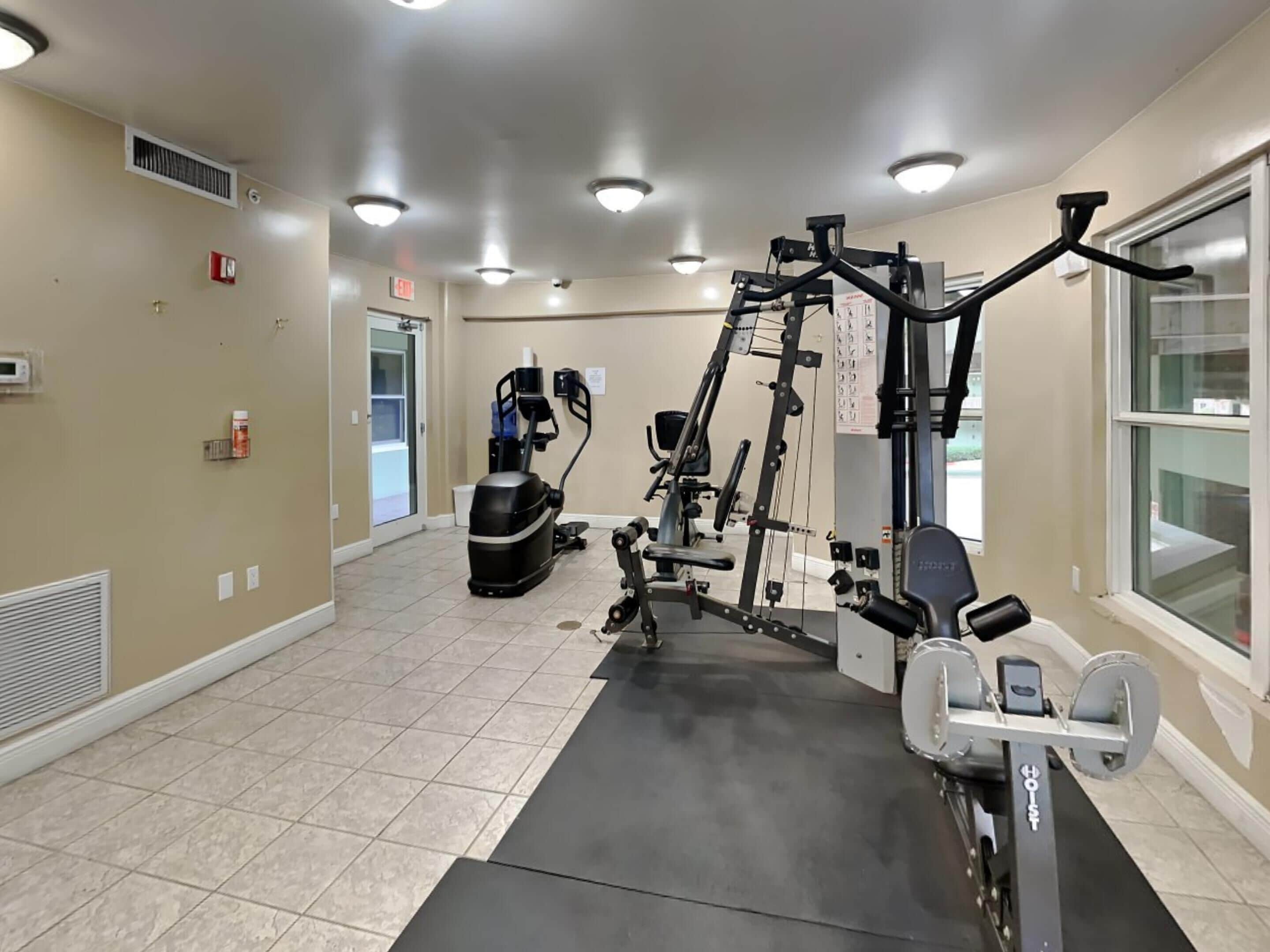 A gym with various exercise equipment, a black mat, and a window on the right.