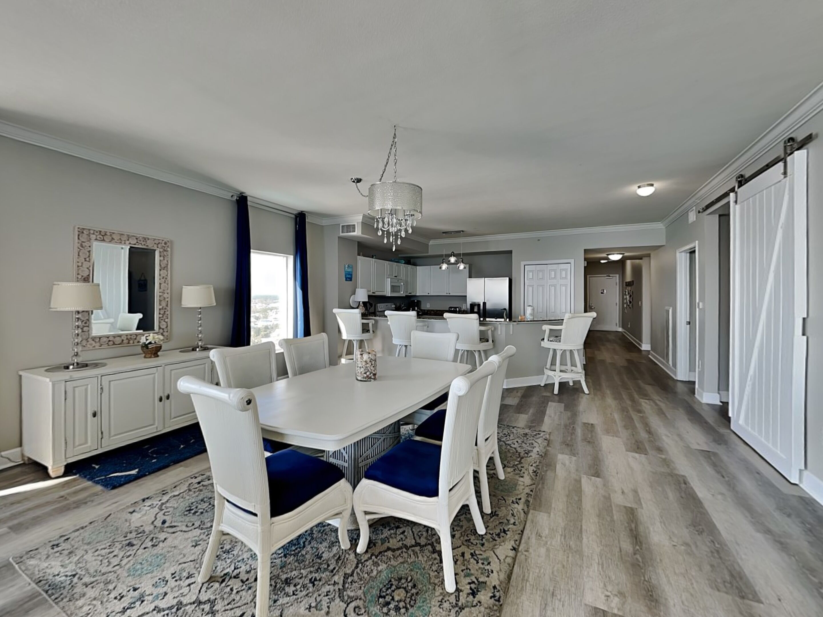 A spacious dining area with a large white table and blue upholstered chairs, a chandelier, and a view of the kitchen in the background.