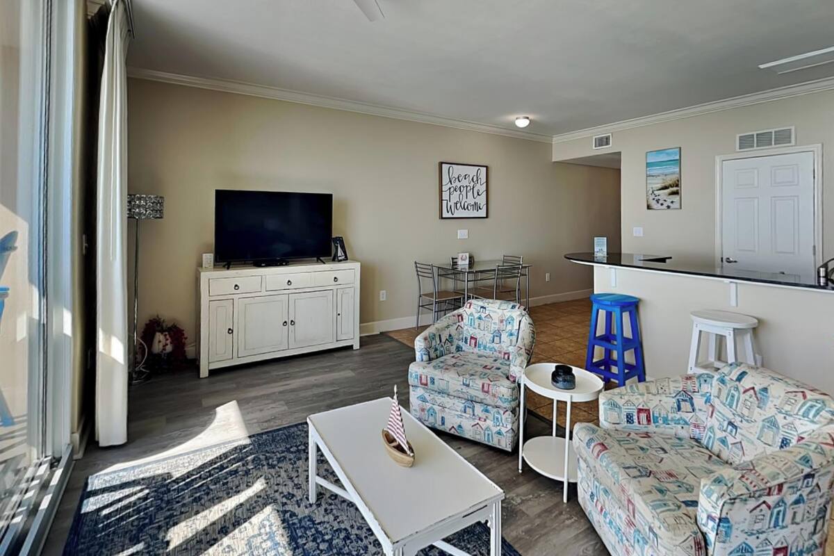 A living room with a TV, sofa, chairs, a coffee table, and a kitchen area with a counter and stools.