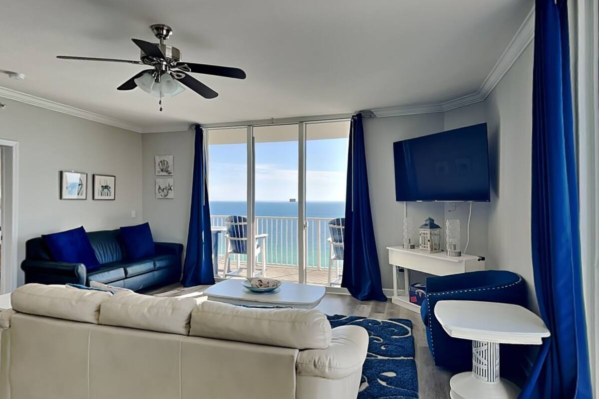 A modern living room with a sofa, a coffee table, and a TV. There is a balcony with a view of the ocean.