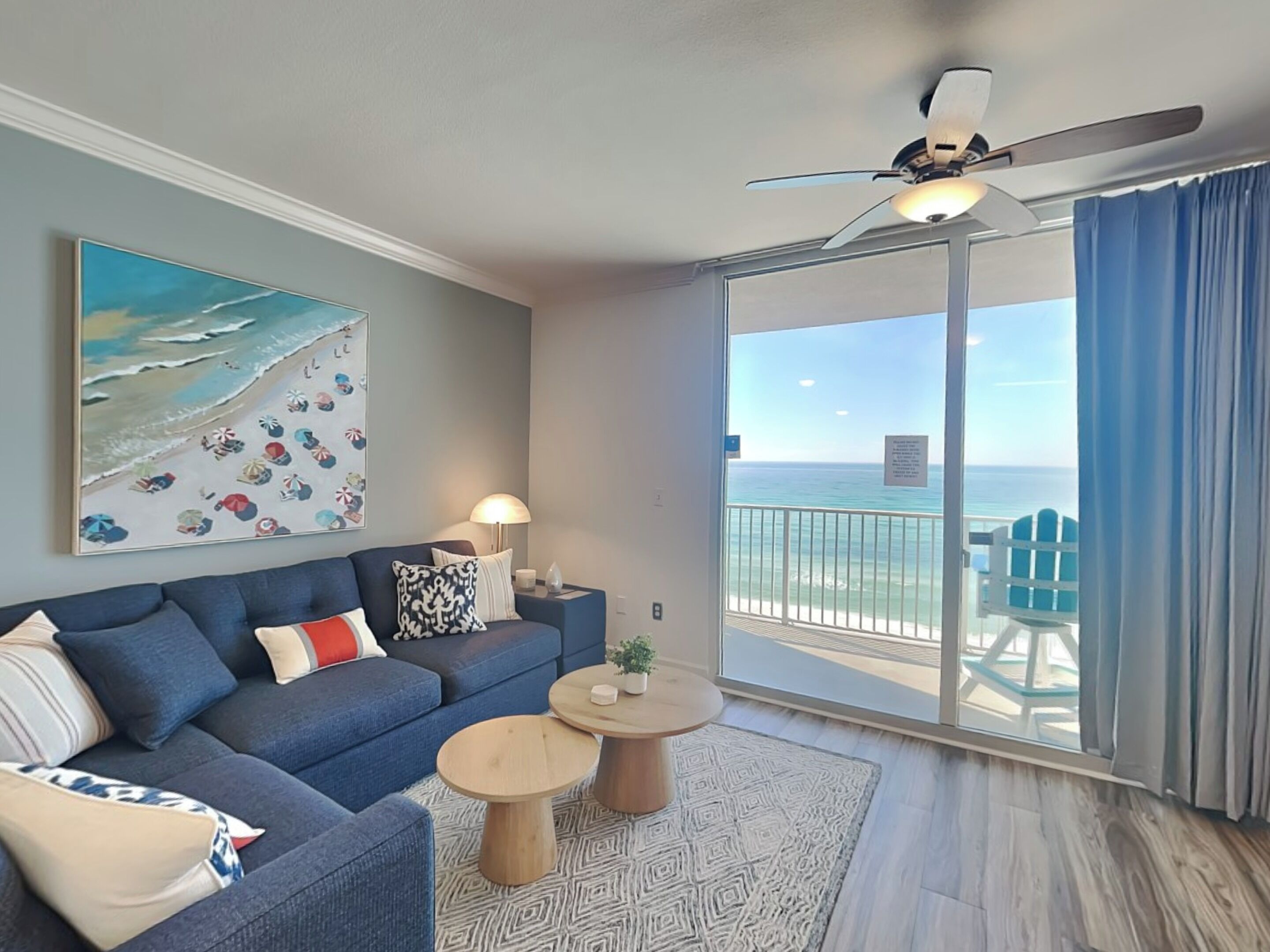 A modern living room with a blue sofa, two round coffee tables, a painting of a beach scene, and a balcony with a view of the ocean.