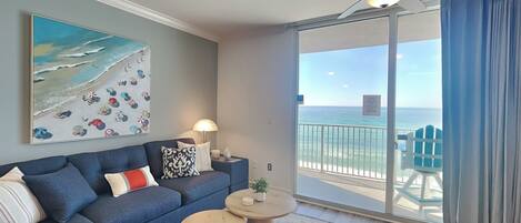 A modern living room with a blue sofa, two round coffee tables, a painting of a beach scene, and a balcony with a view of the ocean.