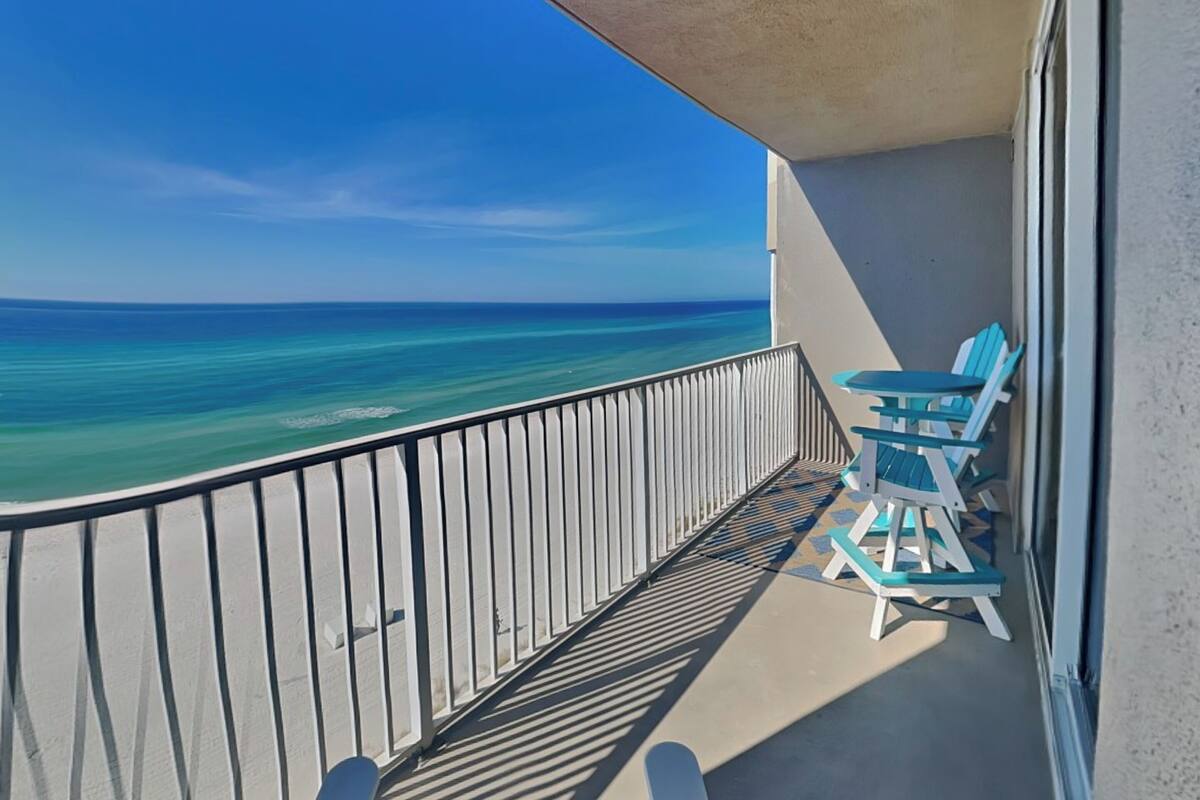 A balcony with a view of the ocean, a white railing, and two turquoise chairs.