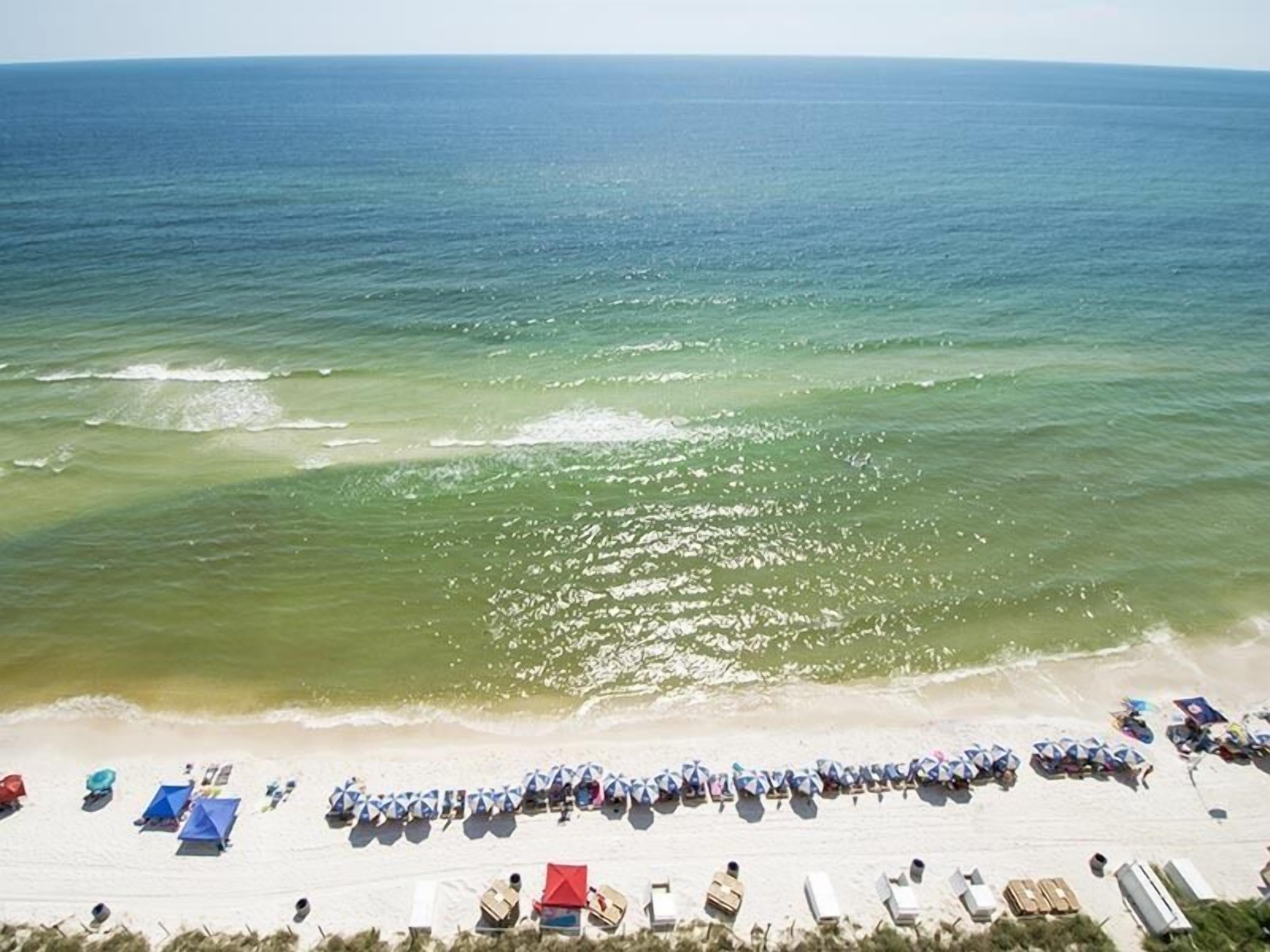 An aerial view of a beach with rows of umbrellas and beach chairs, a sandy shoreline, and the ocean in the background.