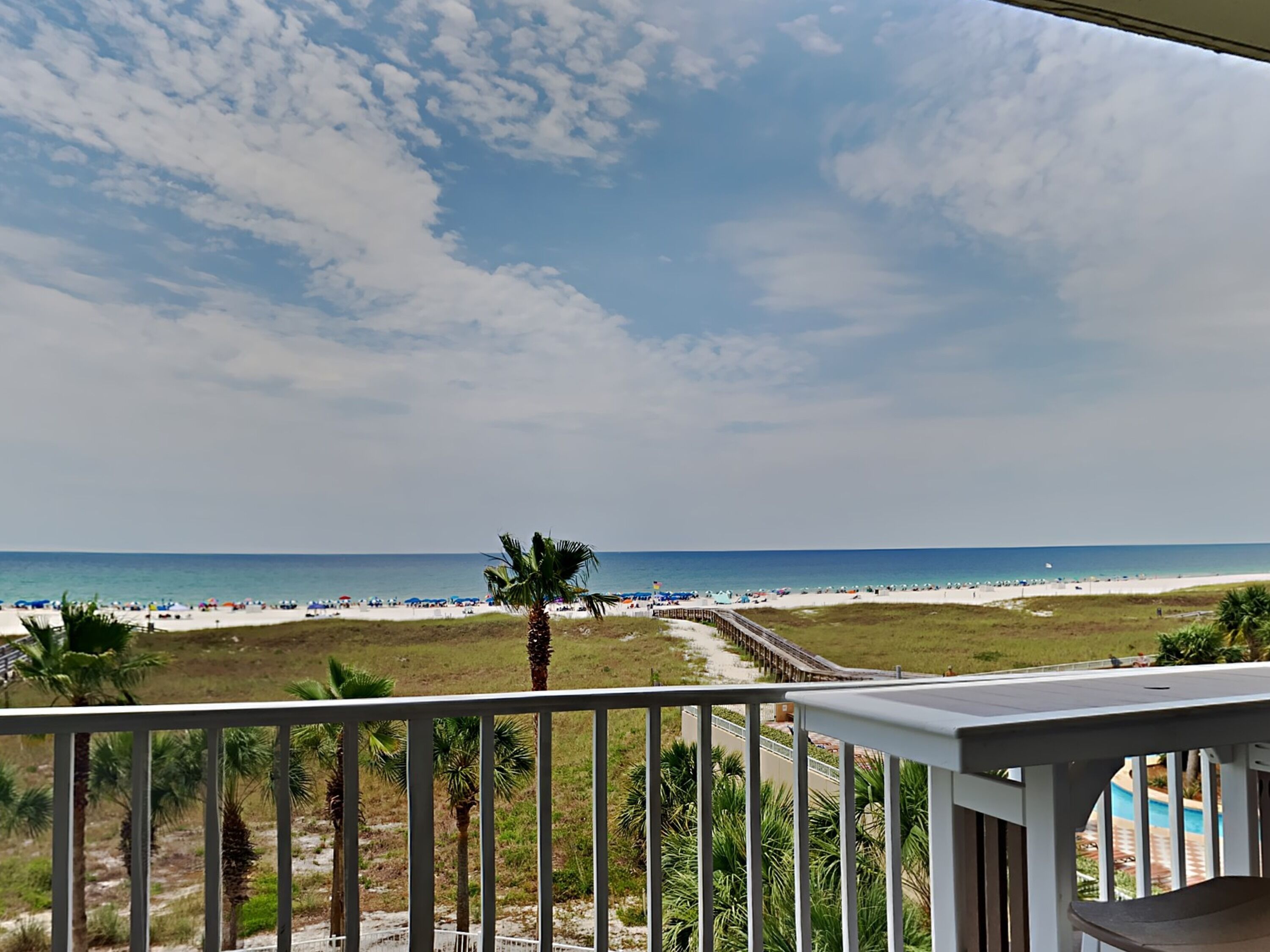A beach view from a balcony with a railing, palm trees, and a clear sky.