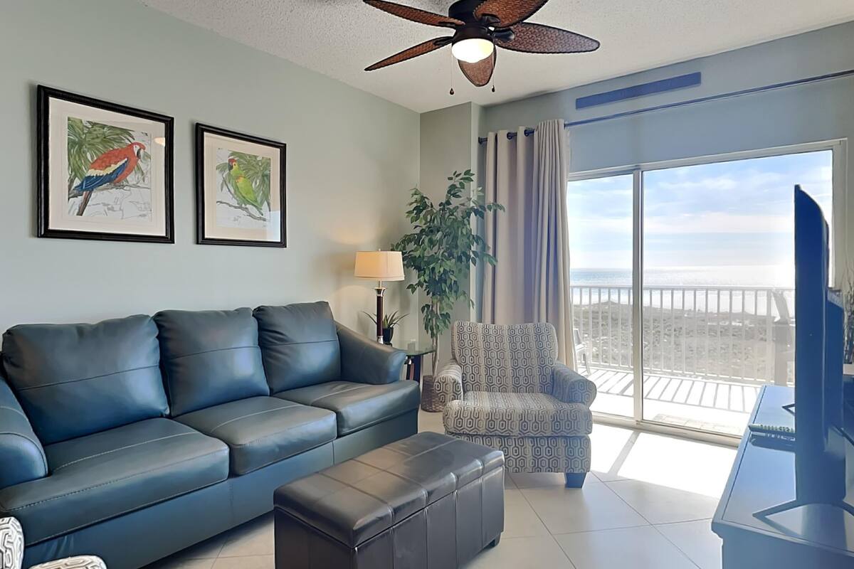 A living room with a blue leather sofa, a patterned armchair, a black ottoman, a ceiling fan, and a large window with a view of the ocean.