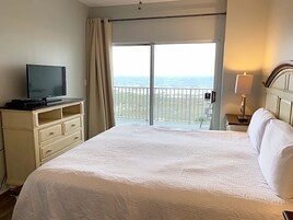 A hotel room with a bed, a television, a nightstand, a lamp, and a balcony with a view of the sea.