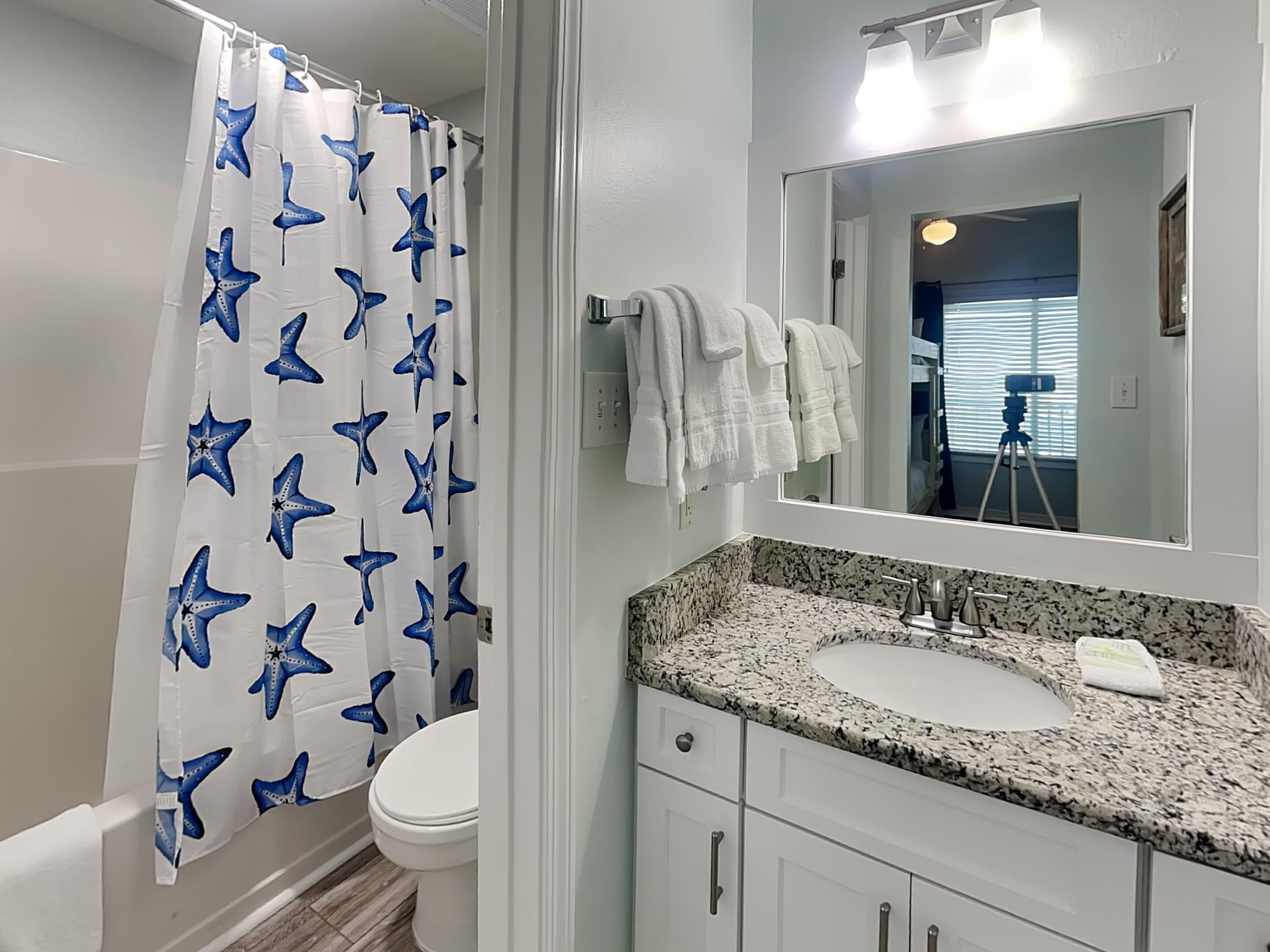 A bathroom with a white toilet, a shower curtain with blue starfish patterns, a granite countertop sink, and a mirror above the sink.