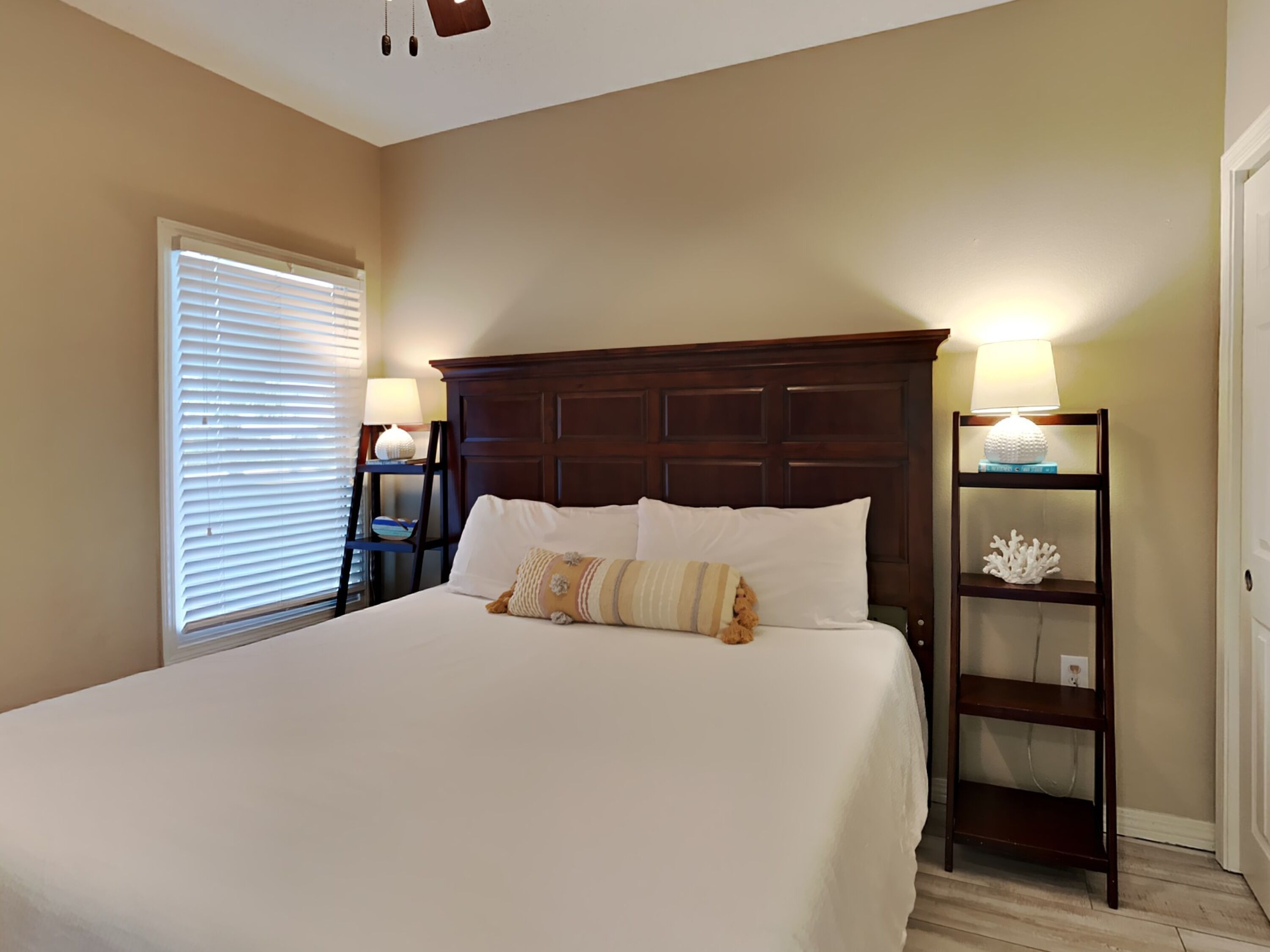 Een netjes opgemaakt bed met witte lakens en decoratieve kussens, een houten hoofdbord, een nachtkastje met een lamp, en een klein plankje met decoratieve objecten.