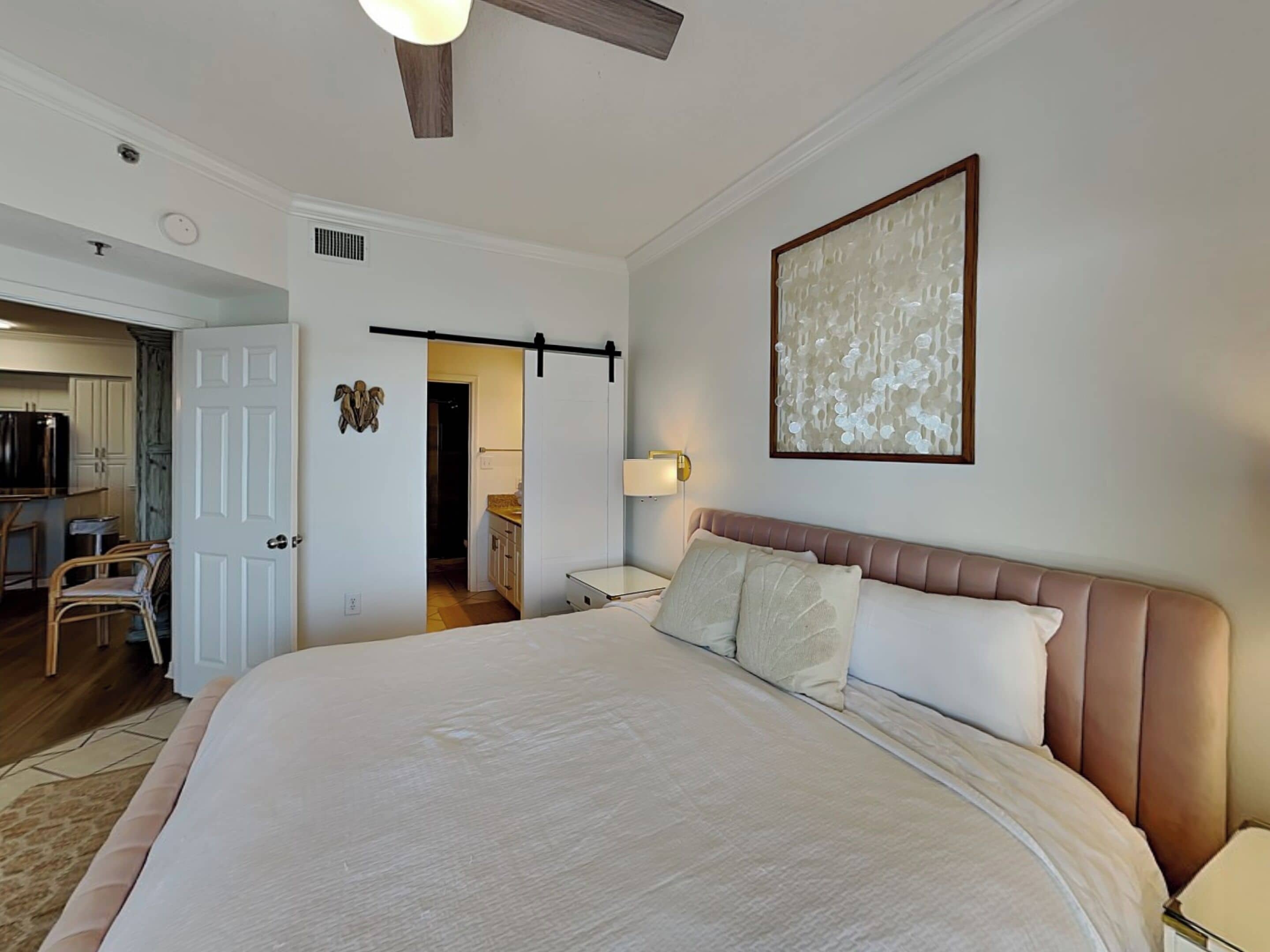 Une chambre à coucher avec un grand lit, une table de chevet, une lampe fixée au mur et une porte donnant sur la cuisine.