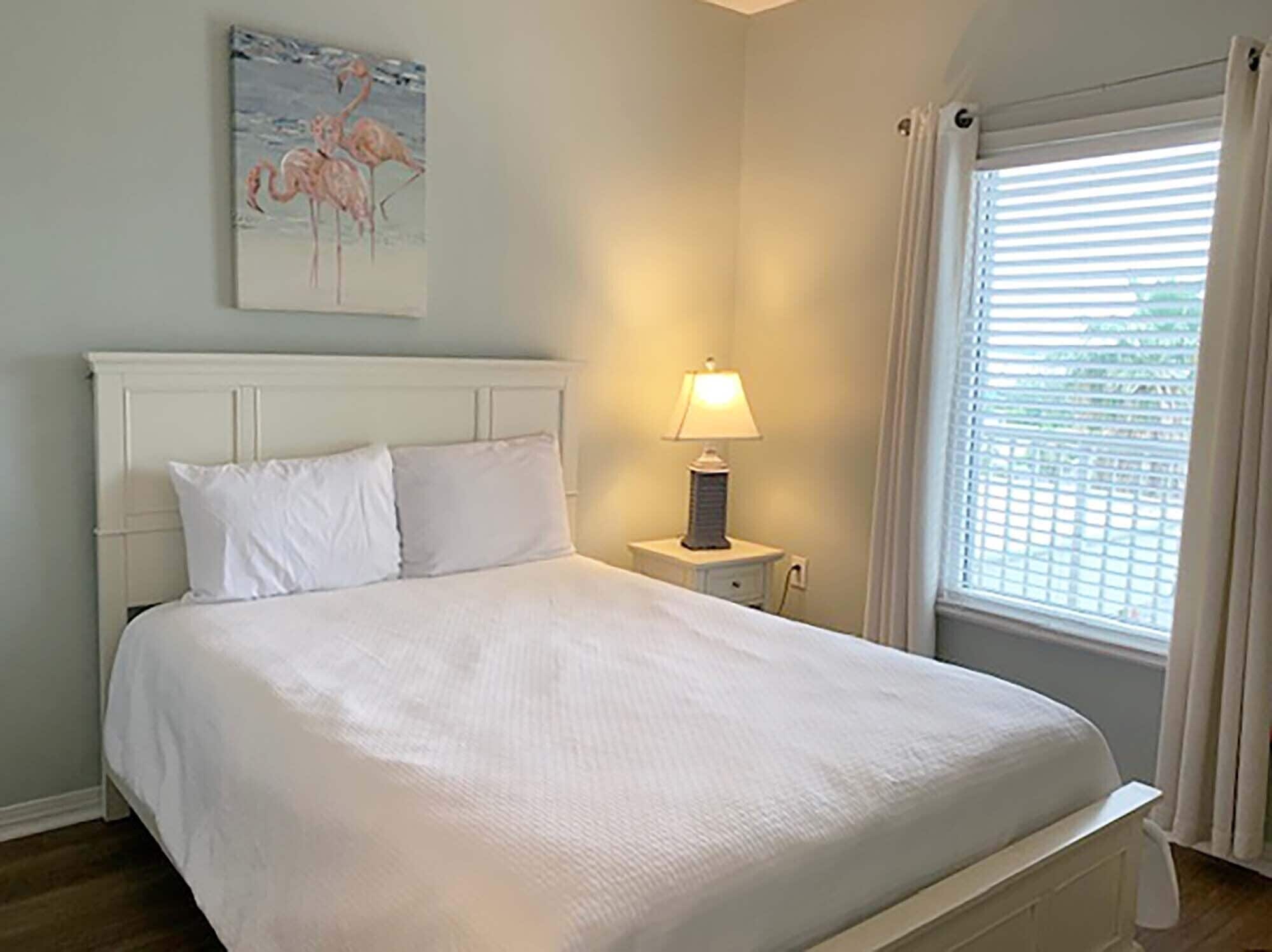 A bedroom with a white bed, a nightstand with a lamp, a painting of flamingos, and a window with blinds.