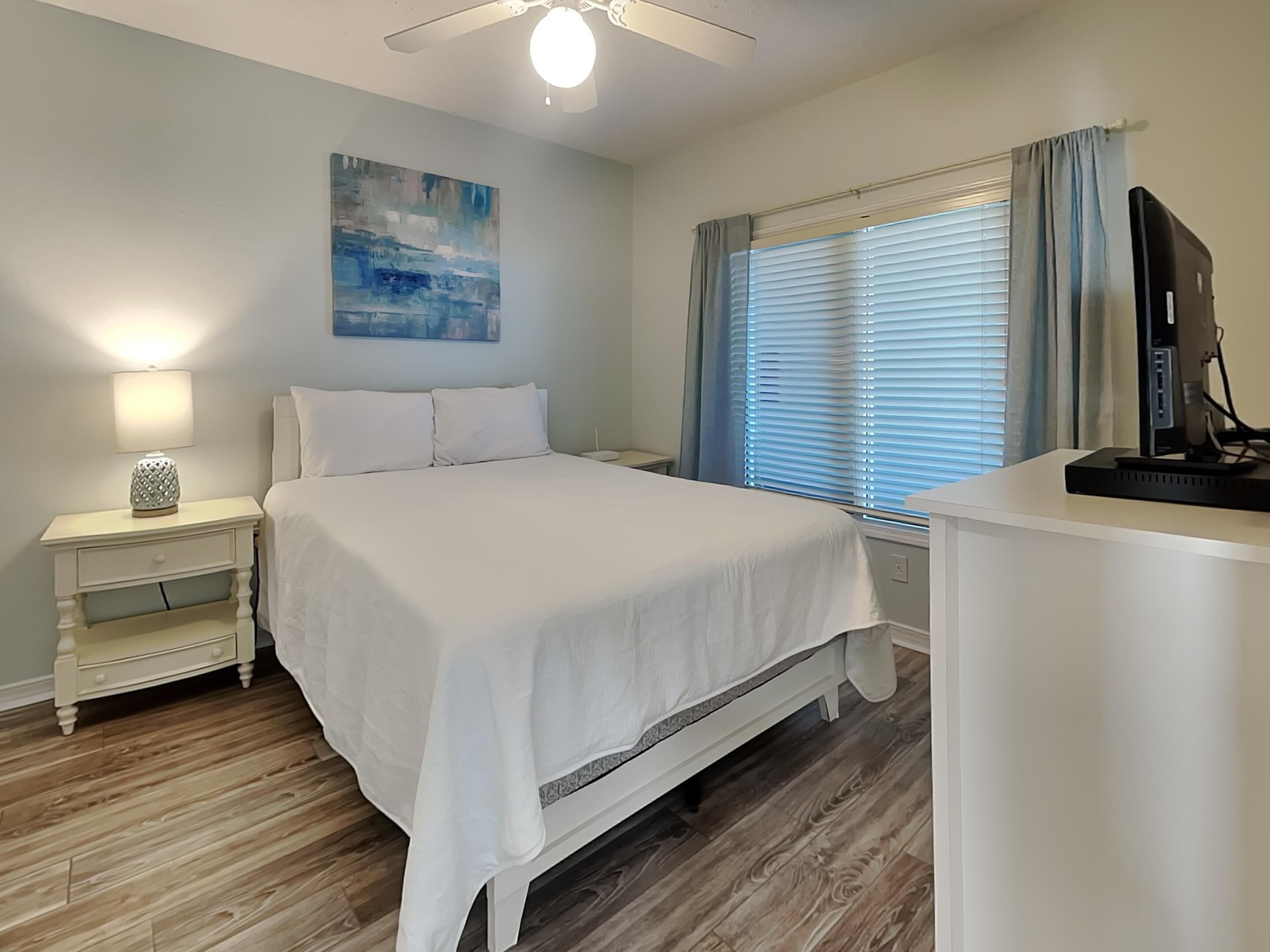 A bedroom with a bed, a nightstand, a desk, a ceiling fan, a window with blinds, and a painting on the wall.