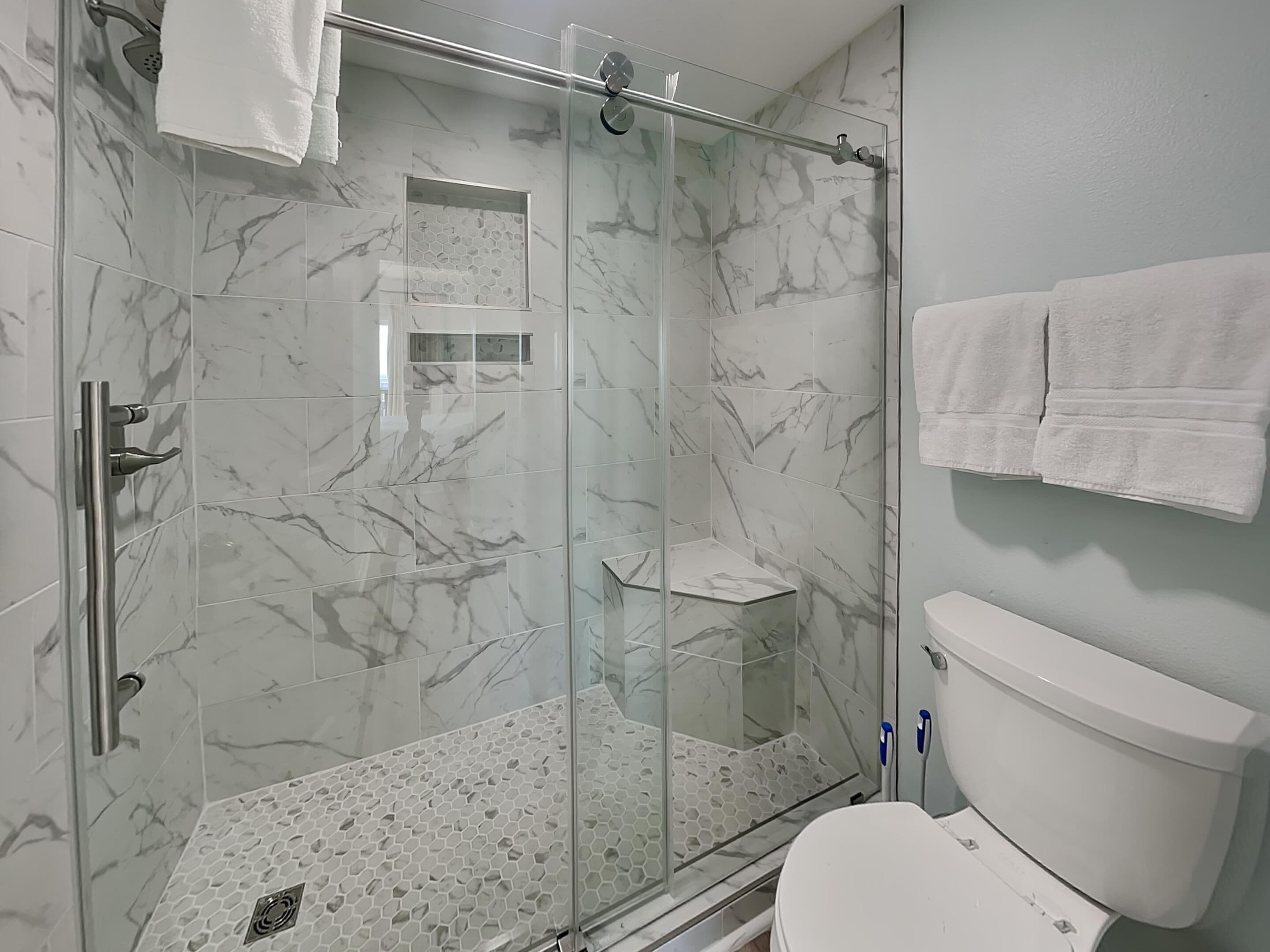 A modern bathroom with a glass-enclosed shower, marble tiles, a toilet, and white towels.