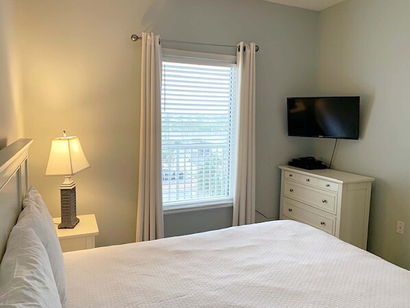 Une chambre à coucher comprenant un lit, une lampe de chevet, une commode, un téléviseur et une fenêtre avec des rideaux.