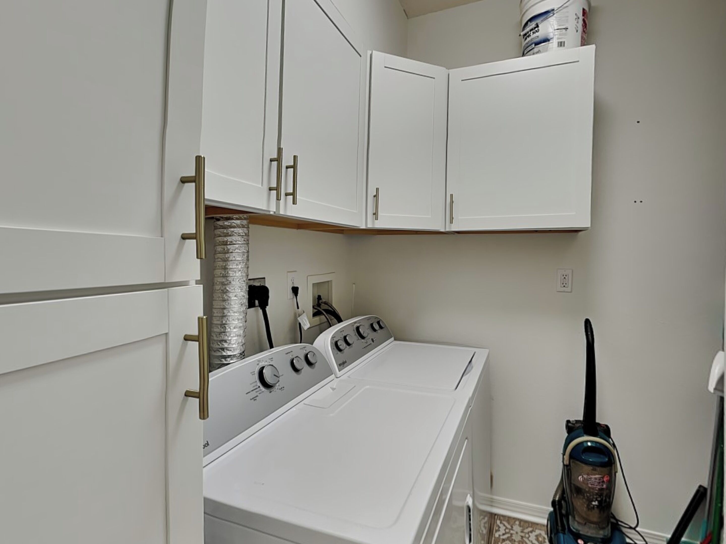 A compact laundry room with a front-loading washer and dryer, white cabinets, and a vacuum cleaner.