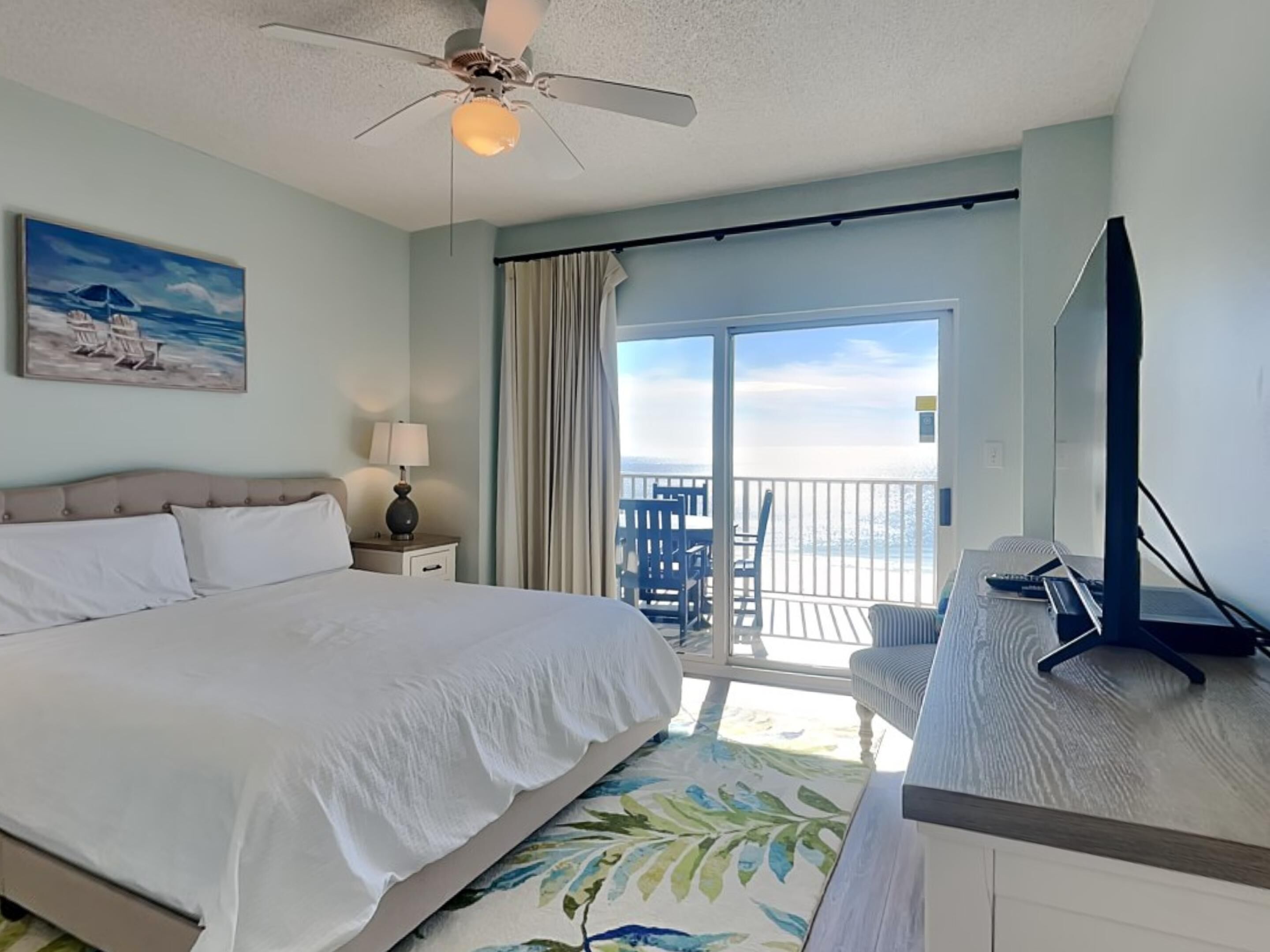 A hotel room with a bed, a desk with a computer, a ceiling fan, a painting, and a balcony with a view of the ocean.