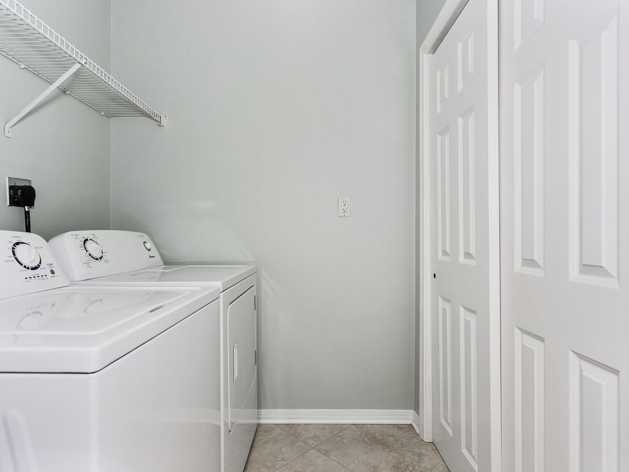 A laundry room with a front-loading washer and dryer, a white door, and a wall-mounted shelf.