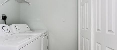 A laundry room with a front-loading washer and dryer, a white door, and a wall-mounted shelf.