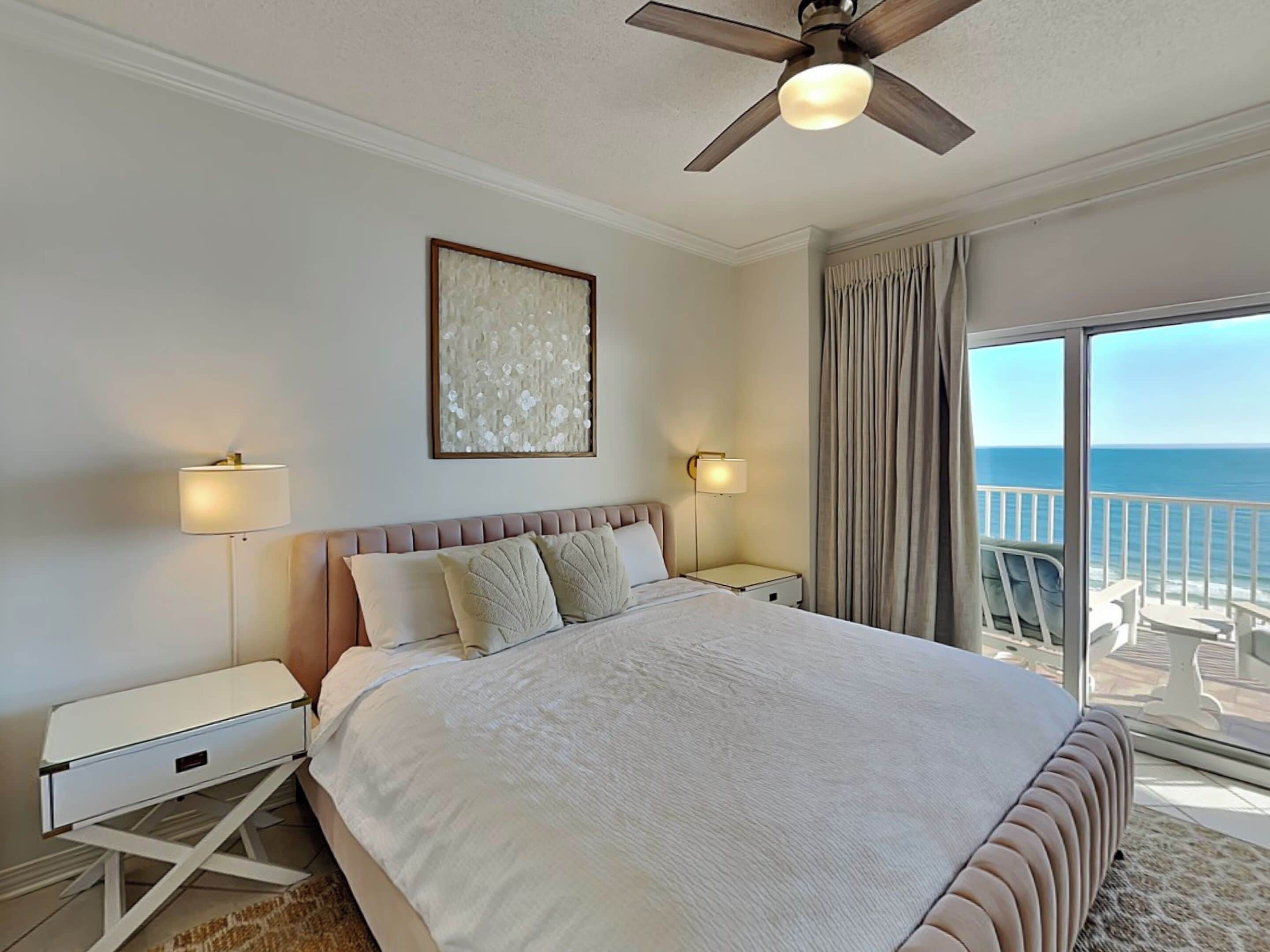 A hotel room with a large bed, two bedside tables, a ceiling fan, a large window with a view of the ocean, and a framed artwork on the wall.