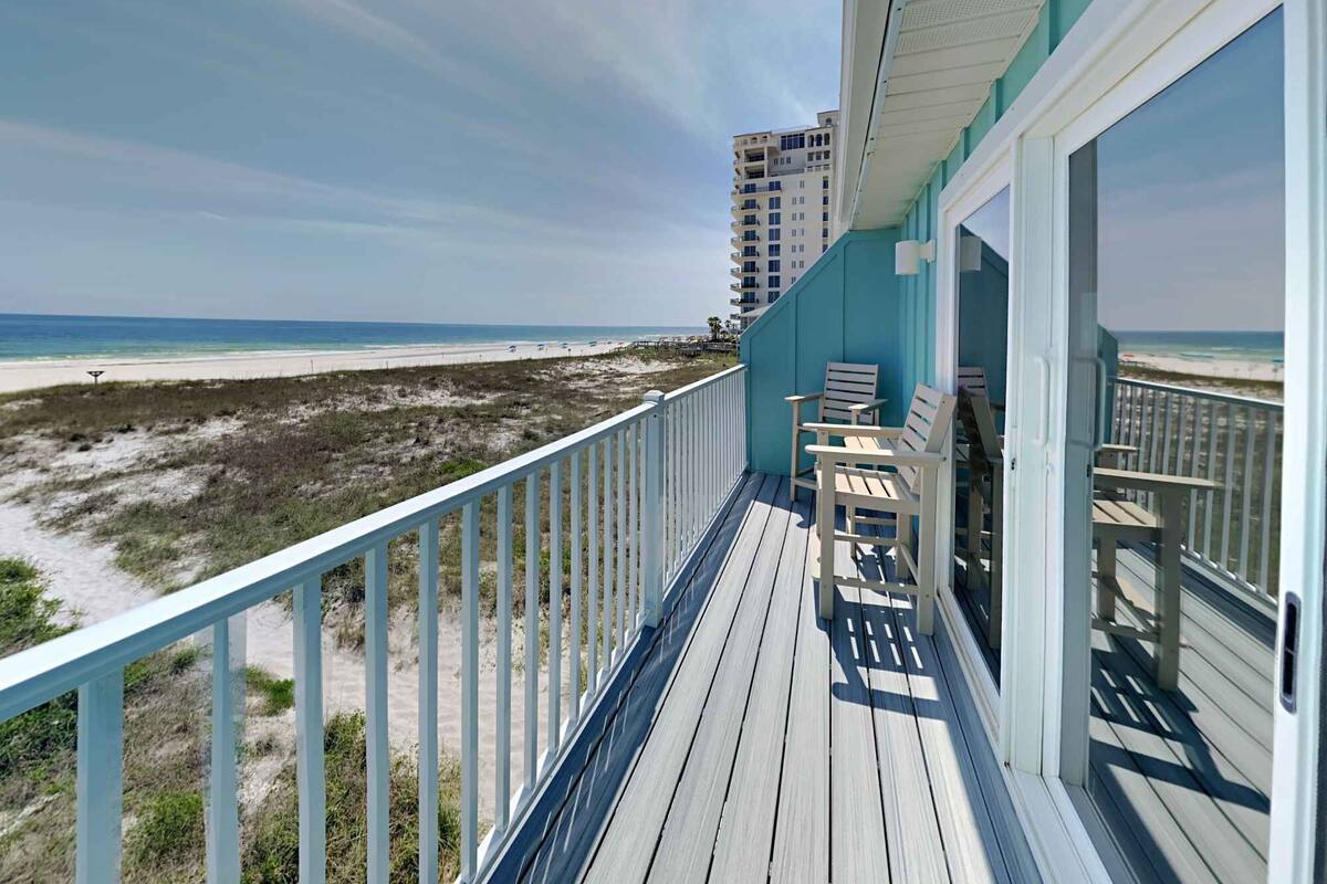 A balcony with a view of the beach and ocean, wooden chairs, and a railing.