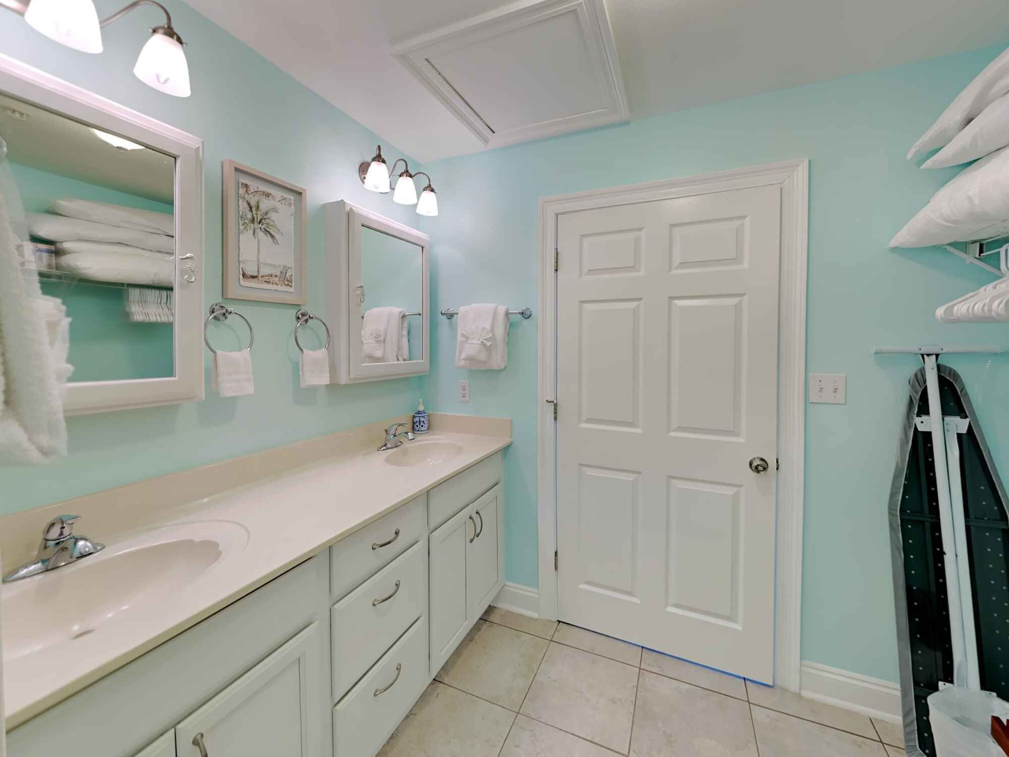 Une salle de bain avec un meuble-lavabo blanc, un évier, un miroir et une porte. Des serviettes sont accrochées au mur, et il y a un porte-serviettes.