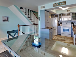 A modern kitchen with white appliances, a dining area with a glass table, and a staircase with a wooden railing.