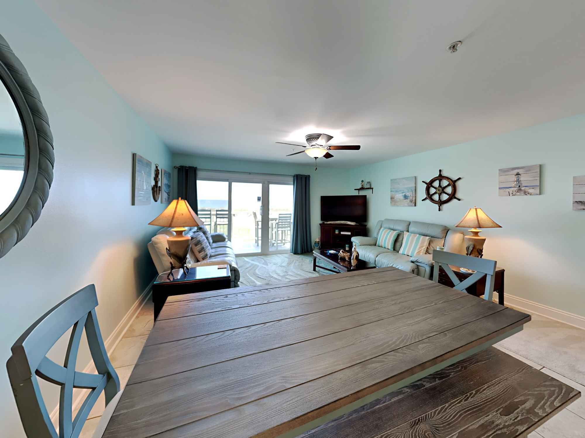 A living room with a large wooden dining table, a sofa, and a TV. There is a ceiling fan, a window with curtains, and a view of the outdoors.