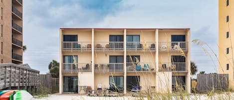A multi-story building with balconies, a sandy foreground, and a sign requesting to keep off dunes.