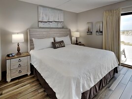 Un dormitorio con una cama grande, dos mesitas de noche con lámparas, un cabecero de madera y vistas a la playa a través de una ventana.
