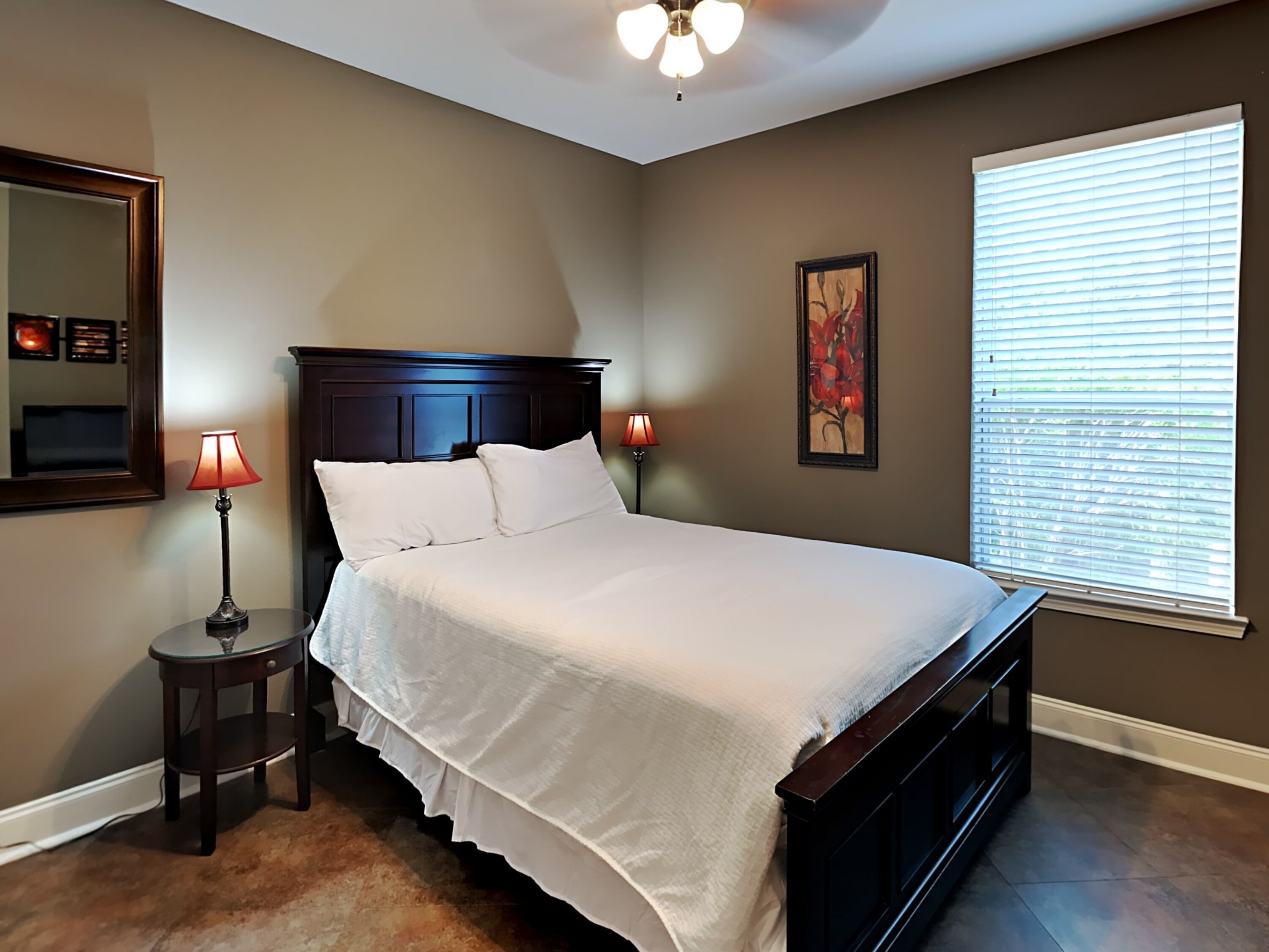 A bedroom with a large bed, two bedside lamps, a ceiling fan, a window with blinds, and a framed picture on the wall.